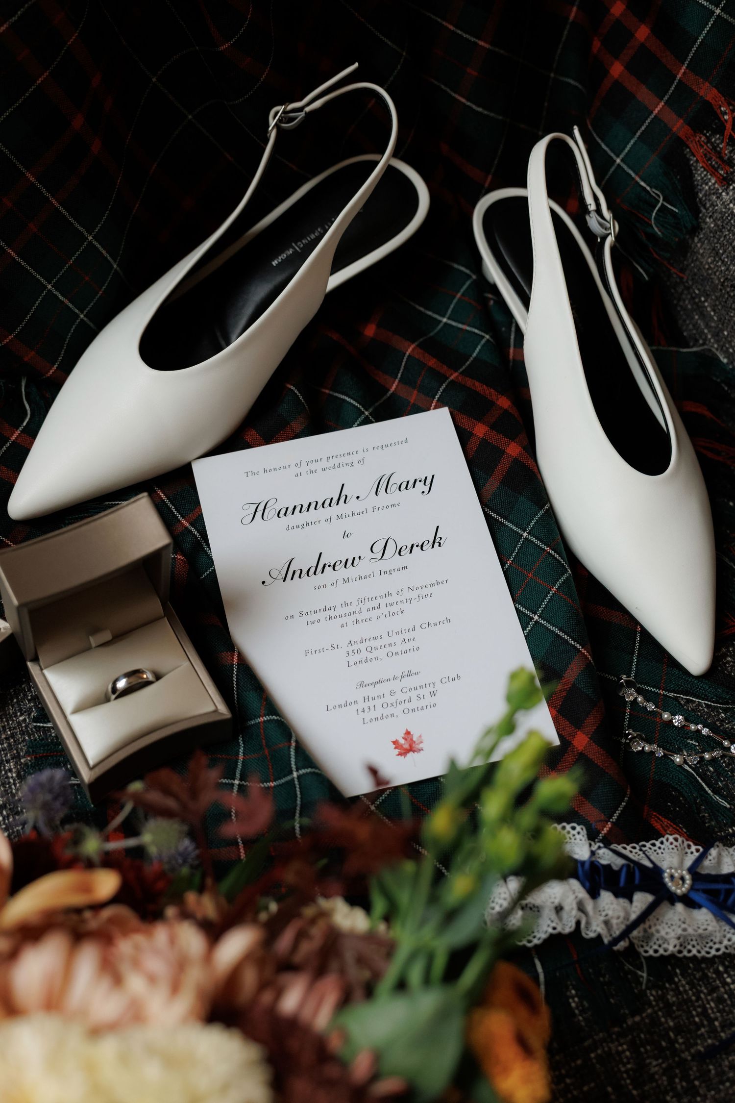 Wedding invitation displayed with white slingback shoes and rings on plaid fabric.