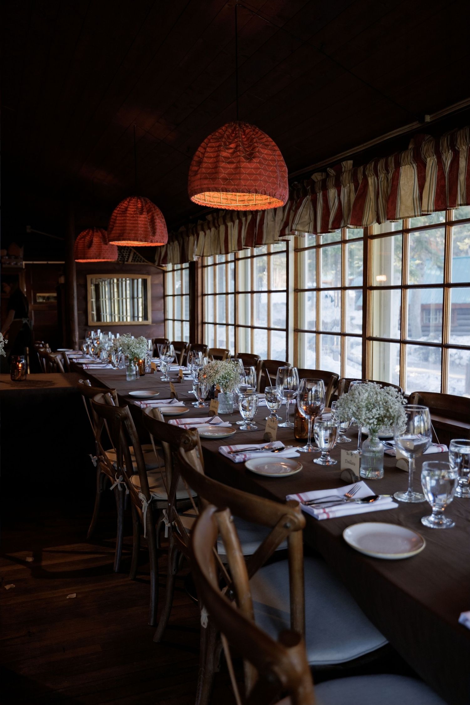 Rustic wedding reception venue with long dining table, cross-back chairs, baby's breath centerpieces, and warm pendant lights.