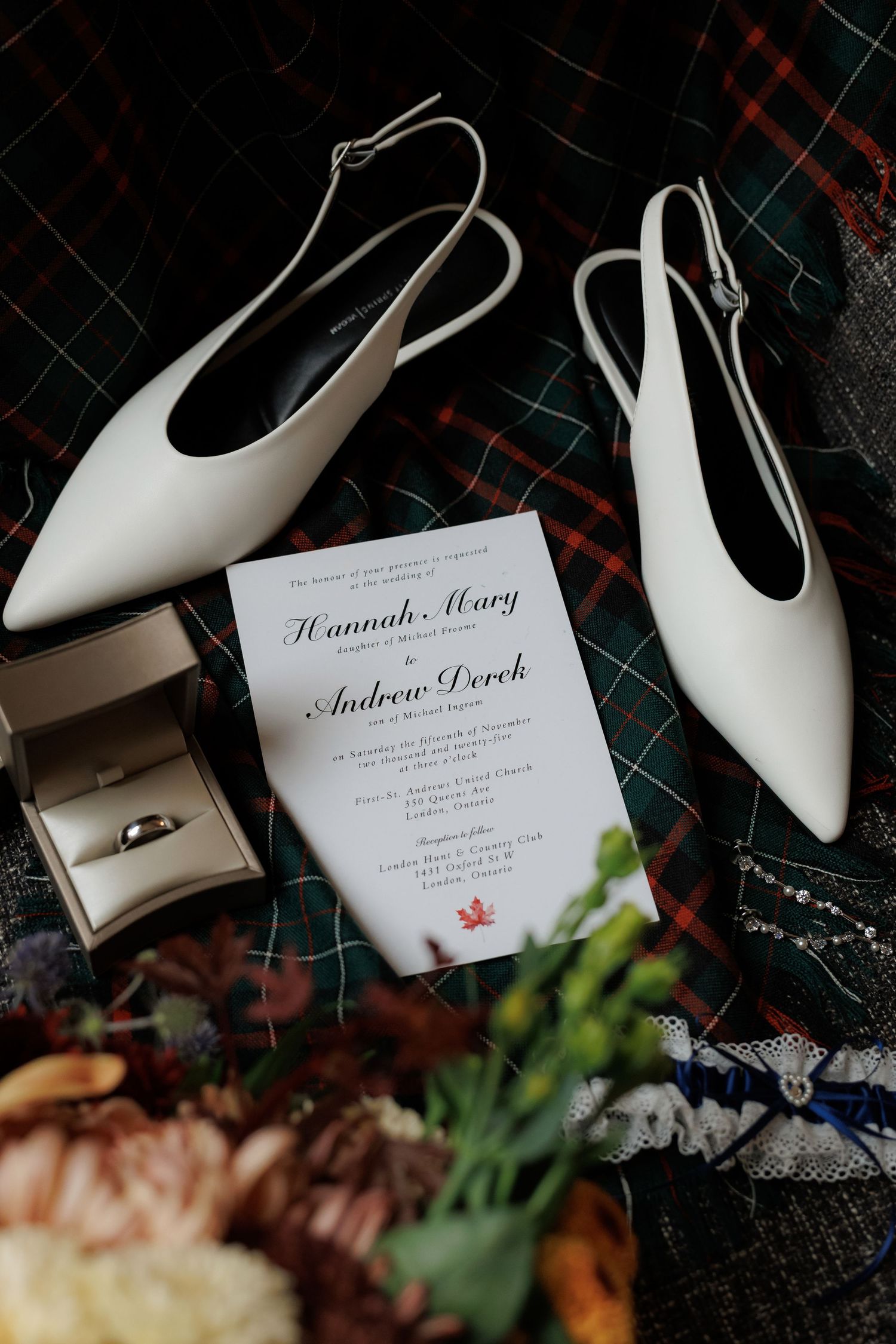 Wedding invitation and white bridal shoes arranged on plaid fabric.