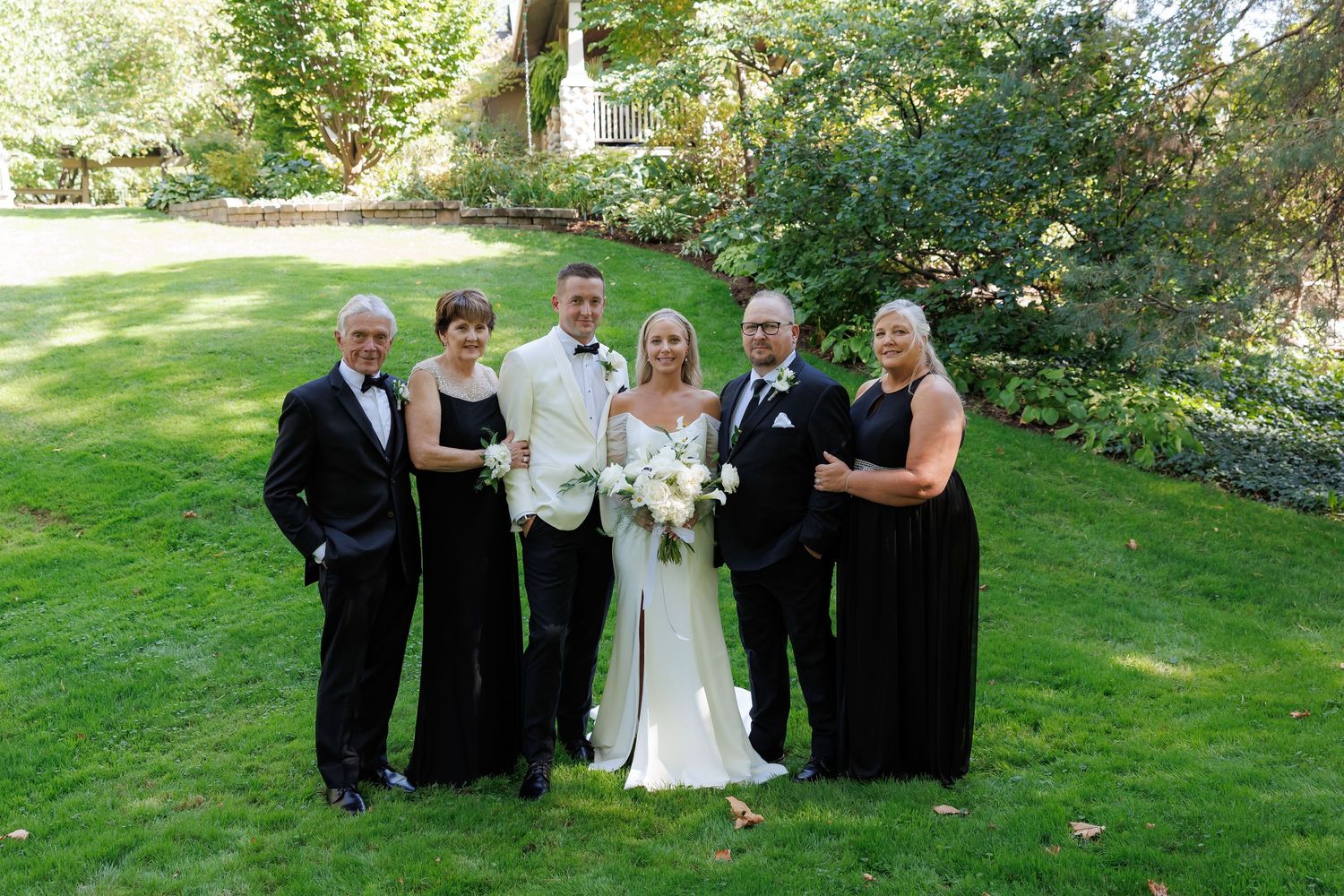 Wedding group photo with bridal party in black and white attire on grassy lawn.