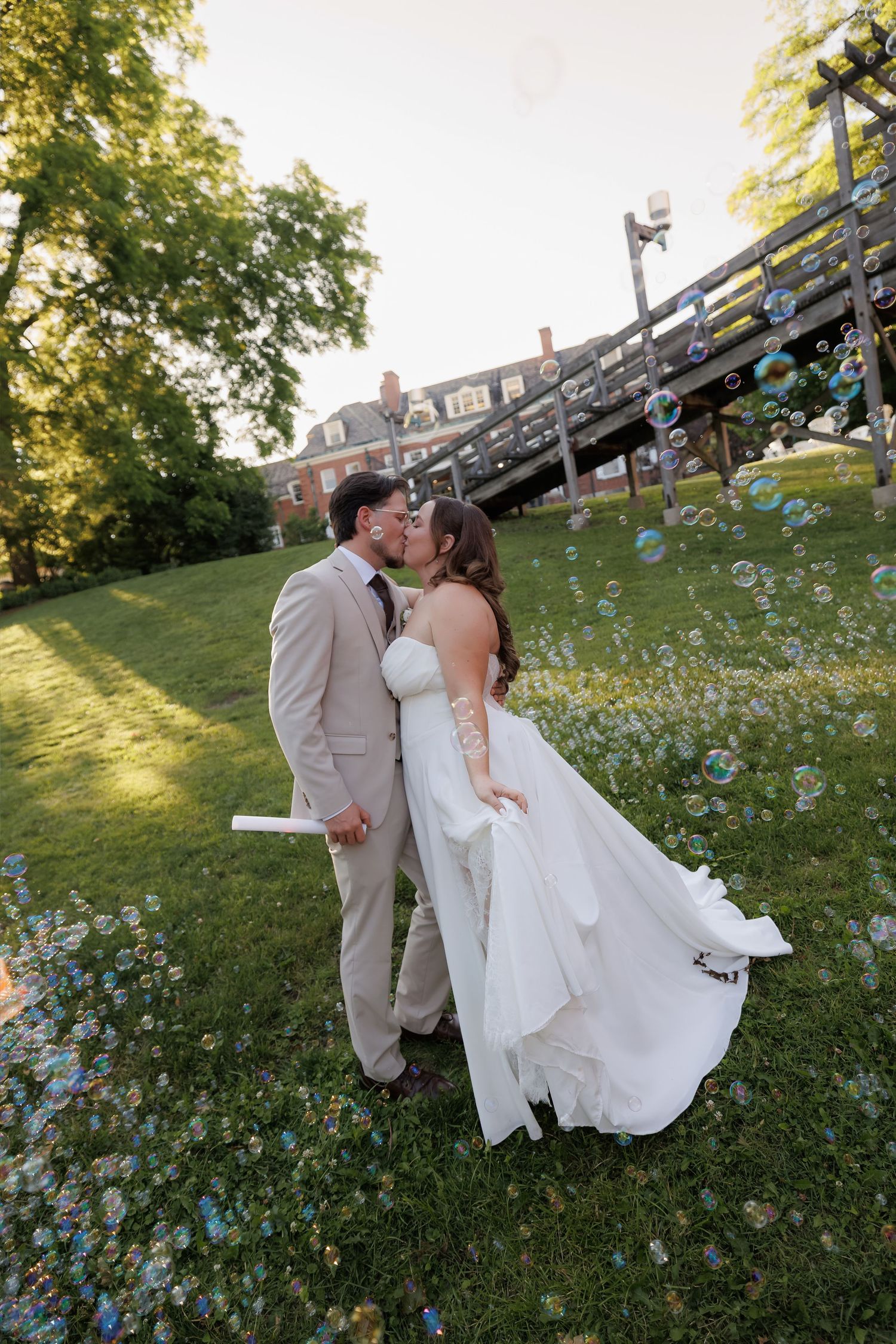 Bride and groom share romantic kiss surrounded by floating bubbles on sunlit green lawn at sunset.