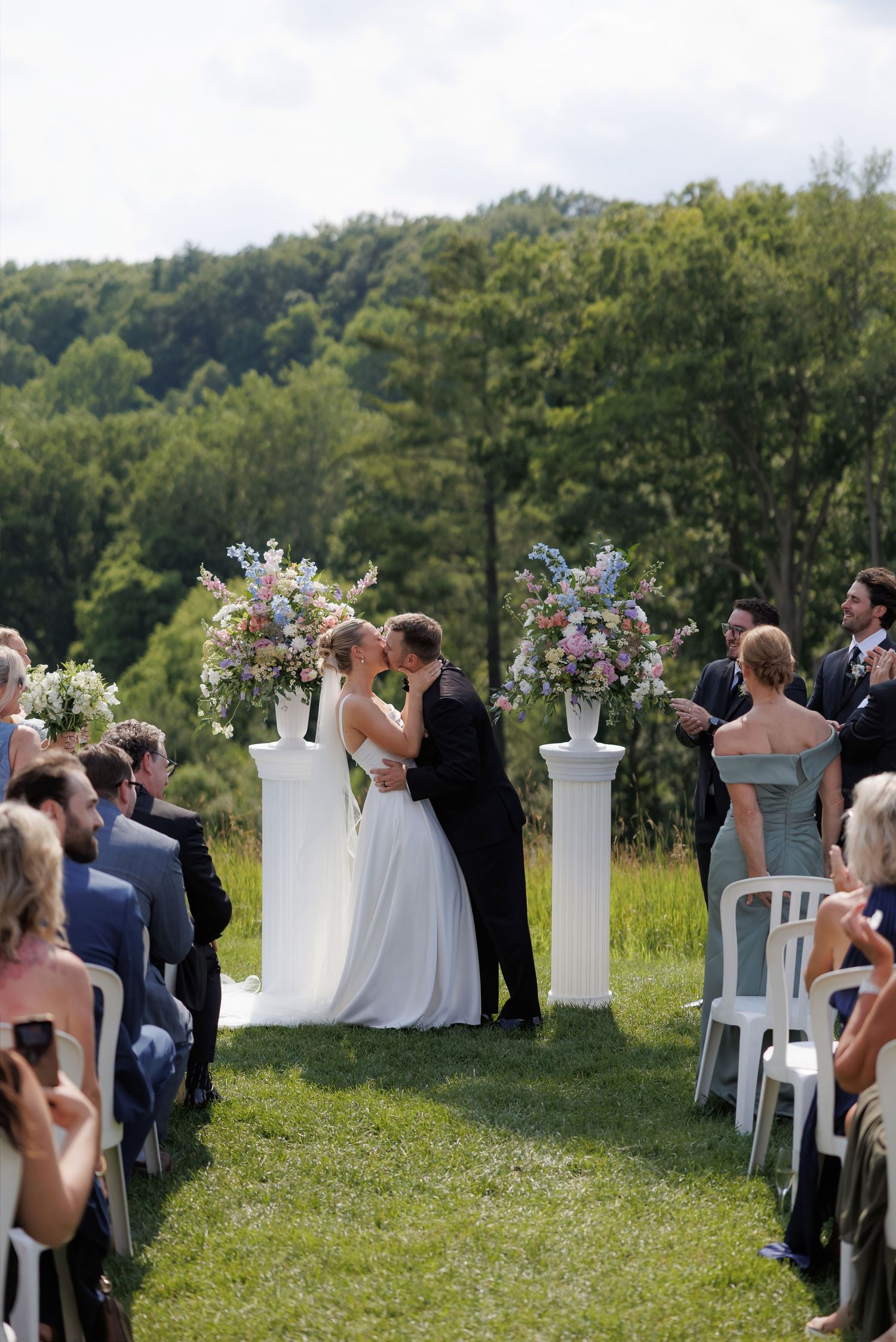 Bride and groom share first kiss at outdoor wedding ceremony with floral arrangements on white pillars.