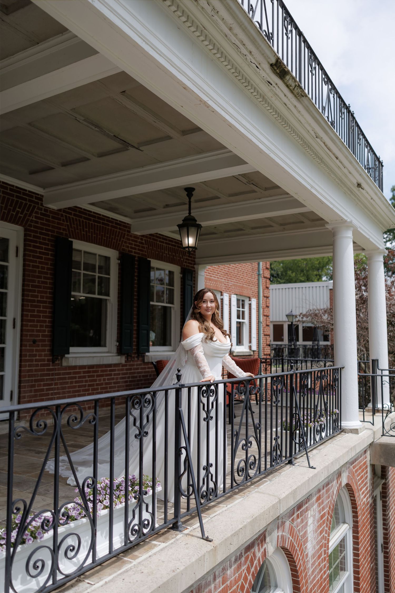 Bride in white off-shoulder gown posing on ornate iron balcony of elegant brick mansion with white columns.