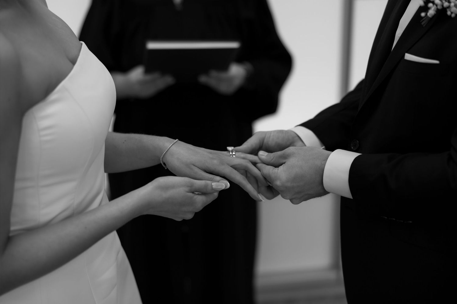 Black and white photo of bride and groom exchanging rings during wedding ceremony with officiant in background.