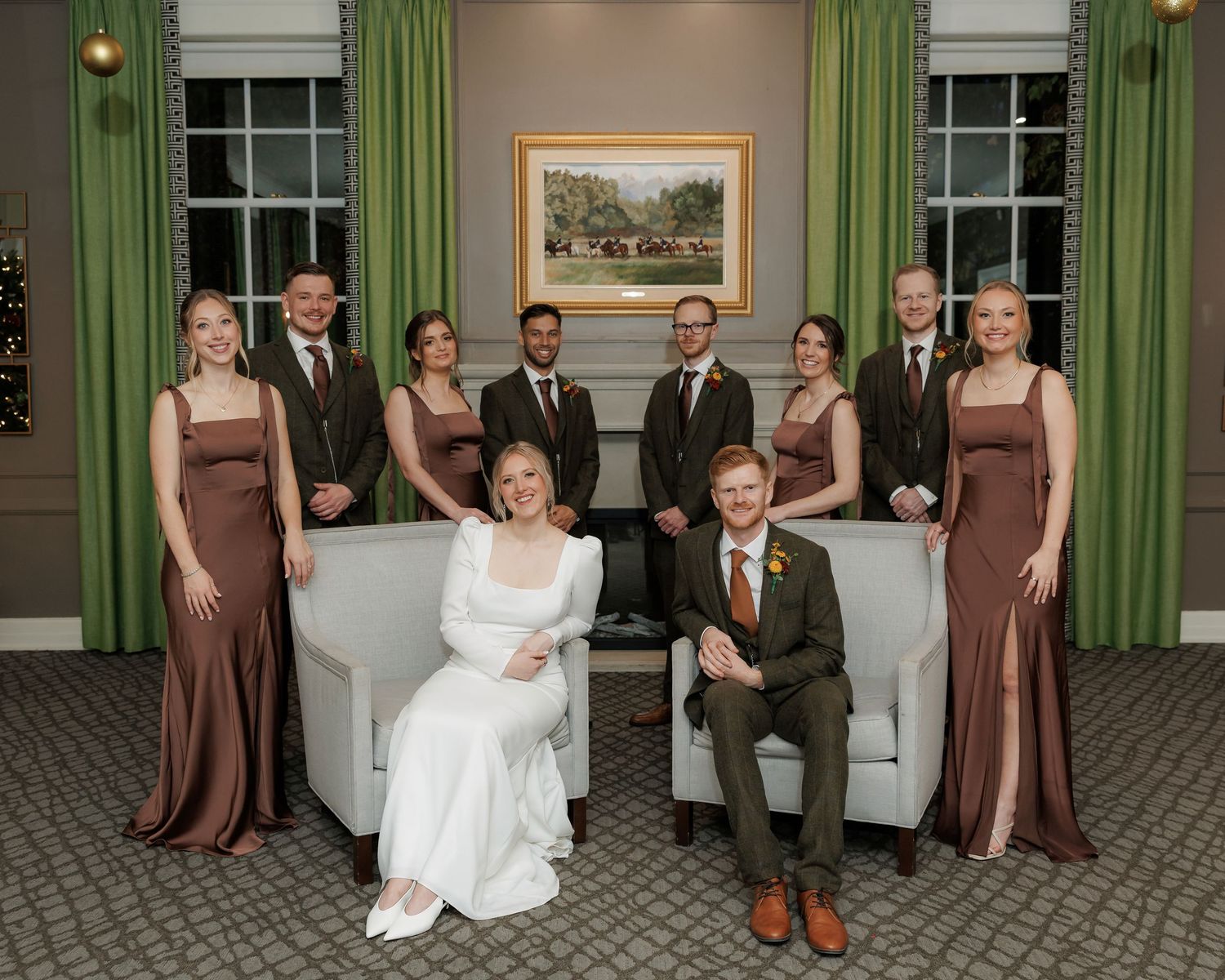 Wedding party members in brown dresses and suits gather for formal photos in an elegant room.