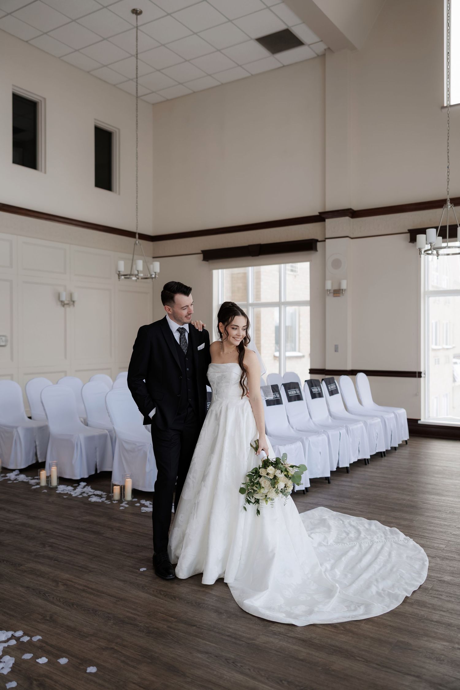 Bride and groom stand together in elegant white ceremony hall with draped chairs and candlelit aisle on their wedding day.