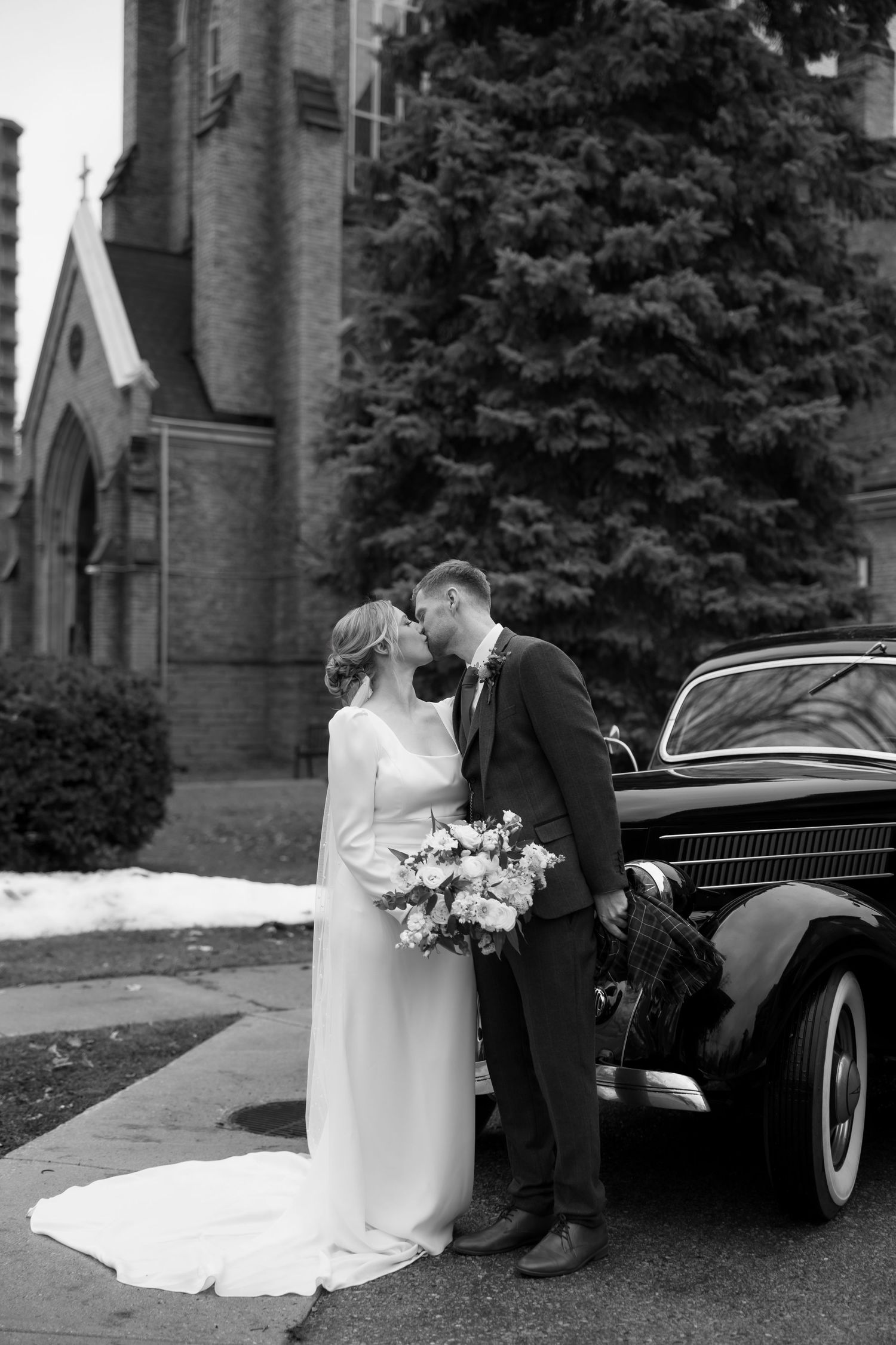 Black and white wedding photos showing a romantic moment next to a vintage car outside a stone church.