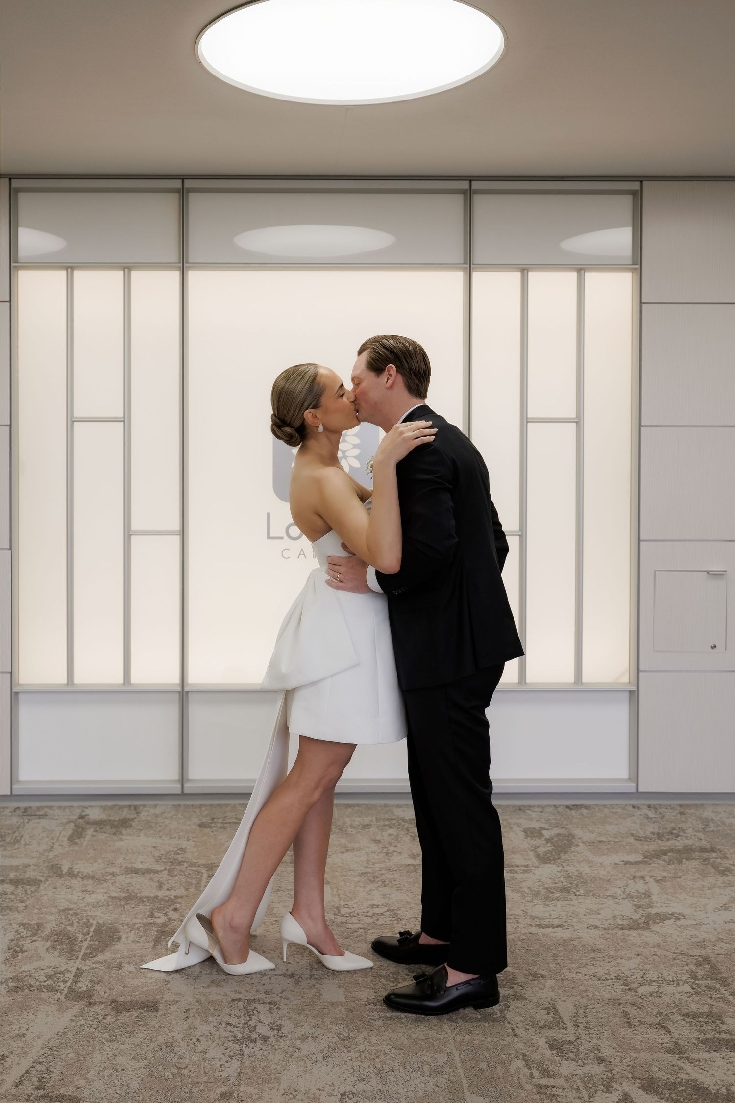 Bride and groom share a first kiss in a modern minimalist interior with soft backlighting behind them.