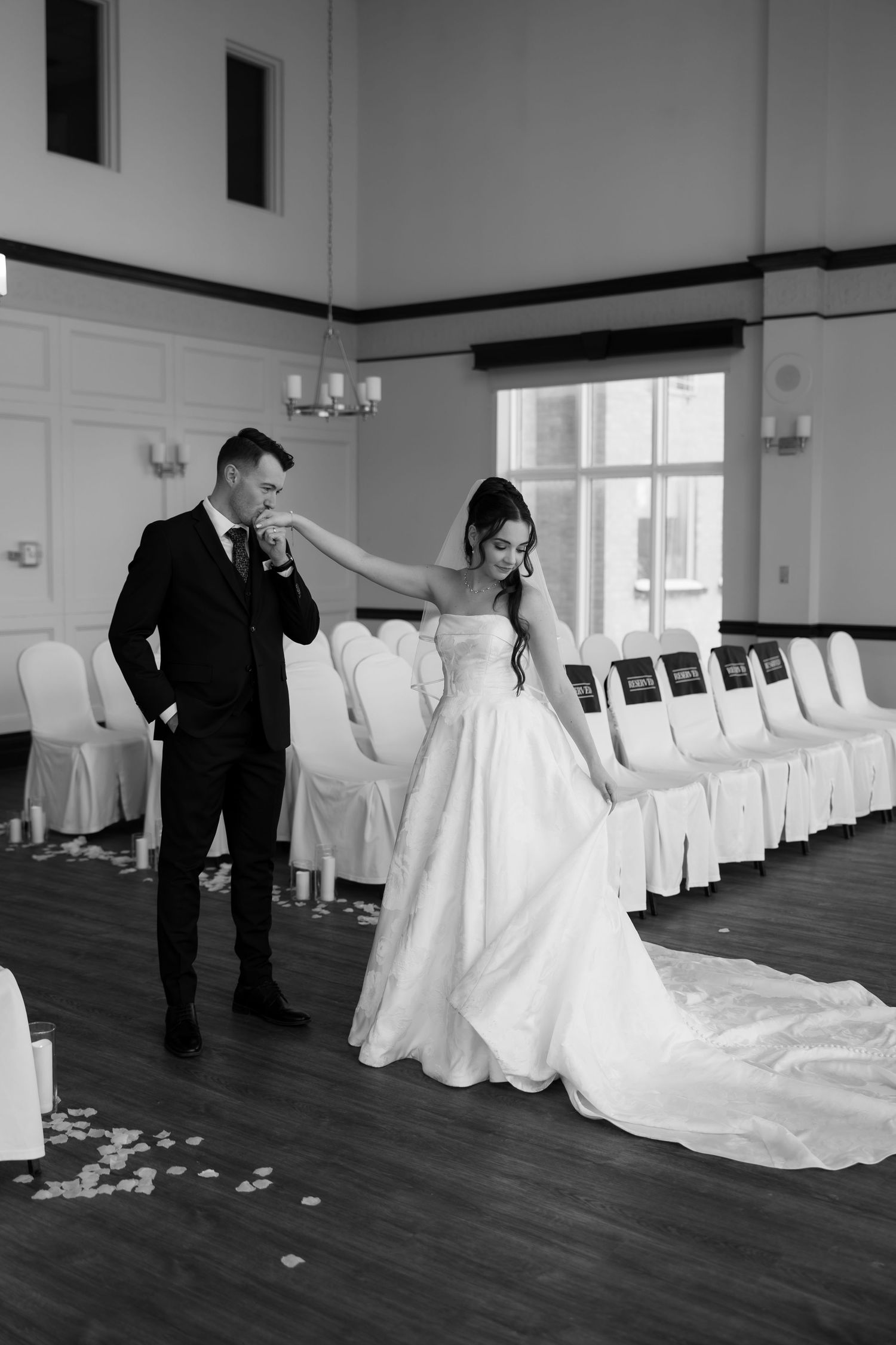 Bride and groom share a tender moment in a wedding venue, groom kissing her hand in black and white.