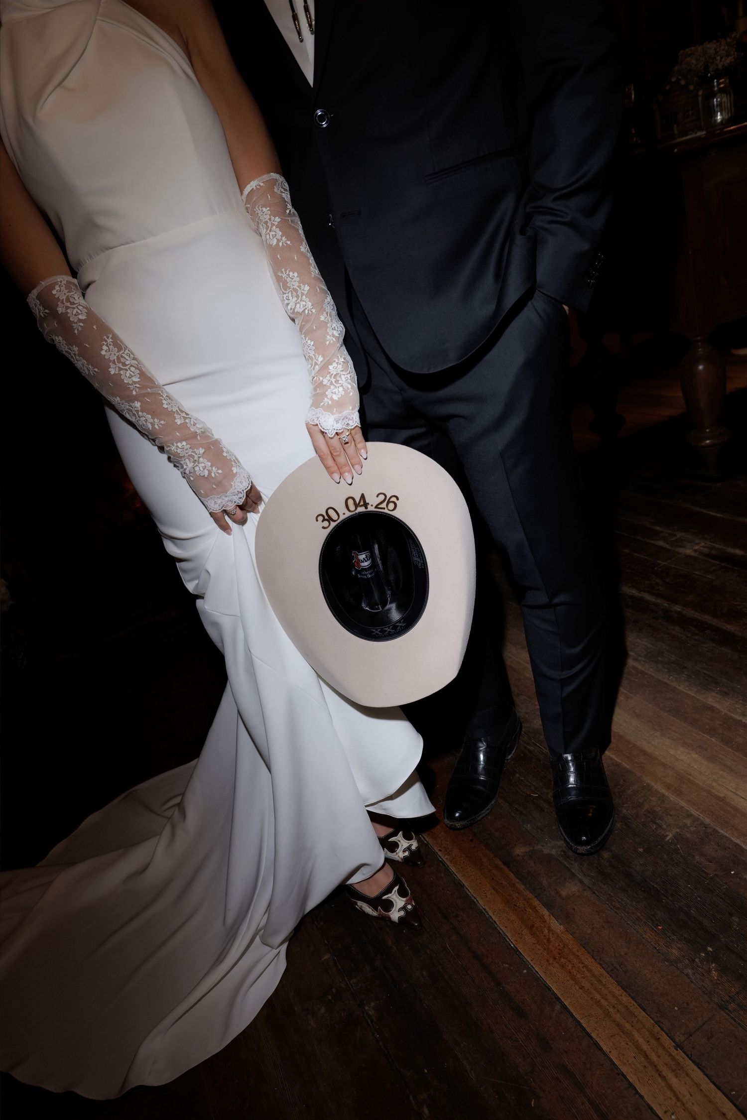 Bride in white gown with lace gloves and groom in black suit holding a wide-brim hat dated 30.04.26 on wooden floor.
