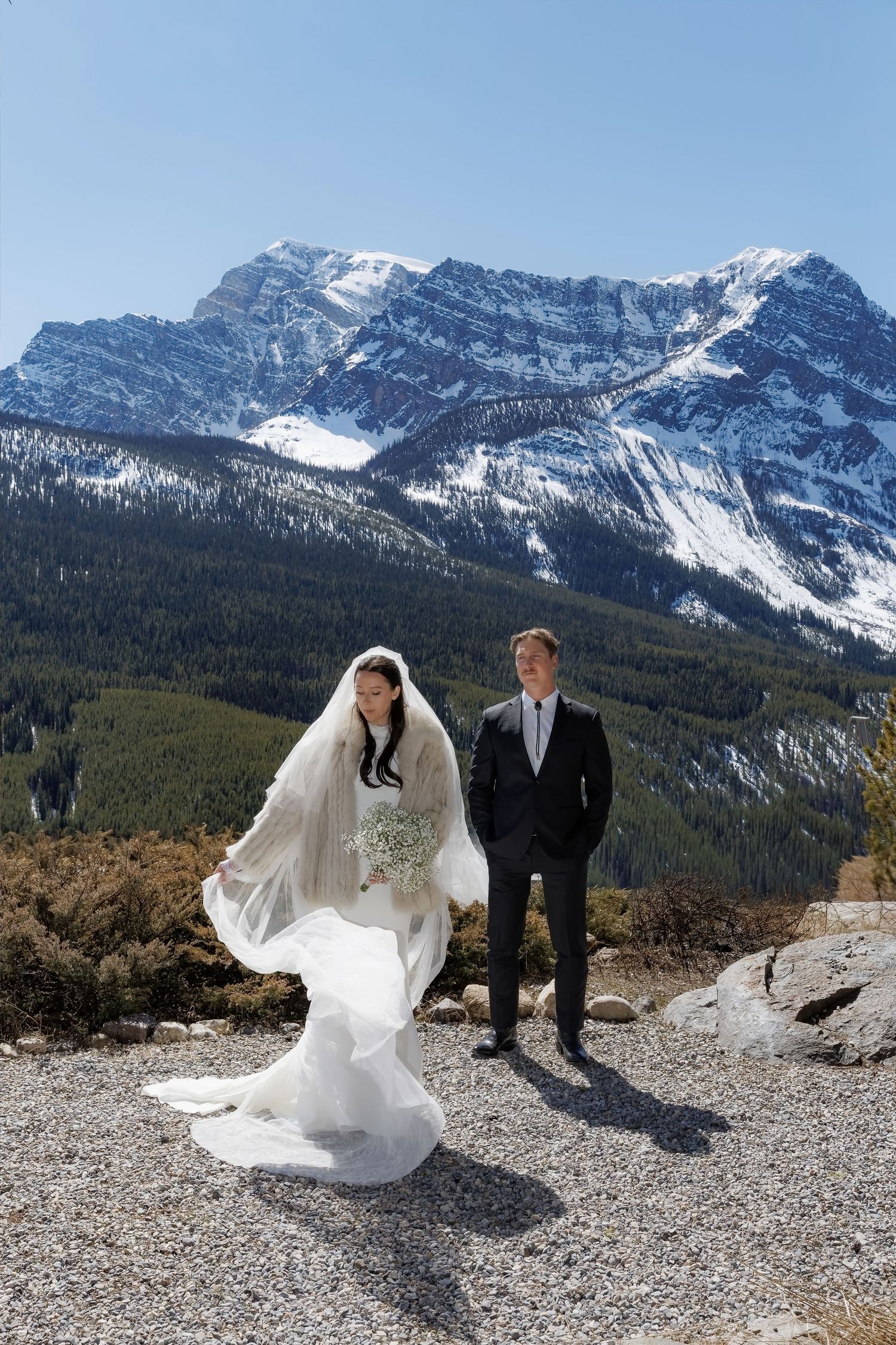 Bride in flowing white veil and groom in black suit pose on gravel with snow-capped Rocky Mountains backdrop.