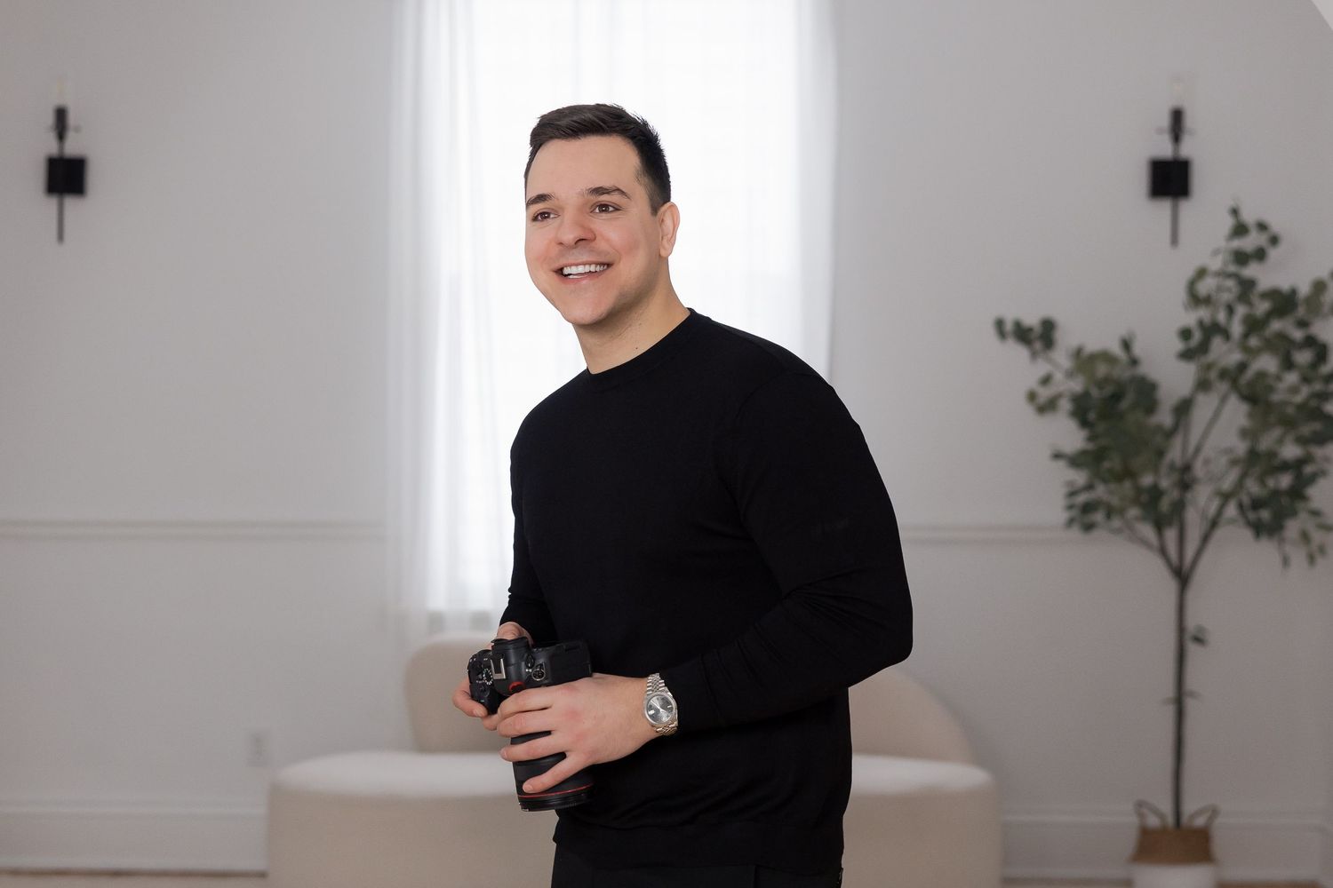 Smiling man in black sweater holding a camera in a bright white studio with a plant and sofa in background.
