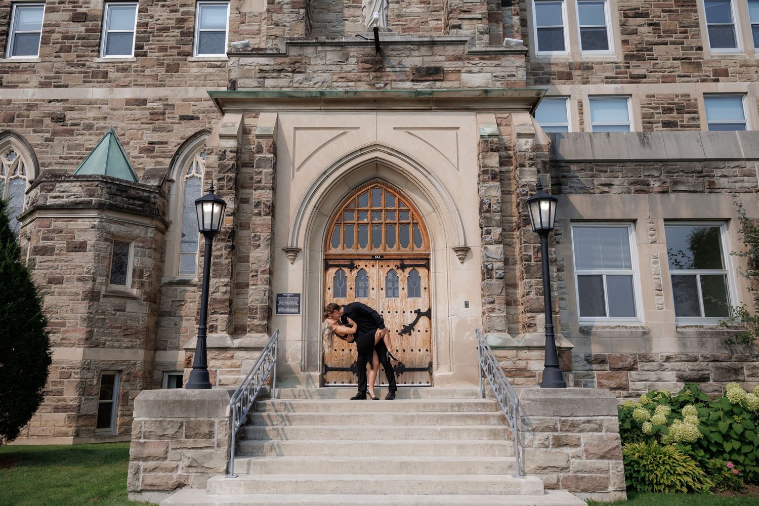A couple stands in the arched stone doorway of a Gothic-style building with stone steps.