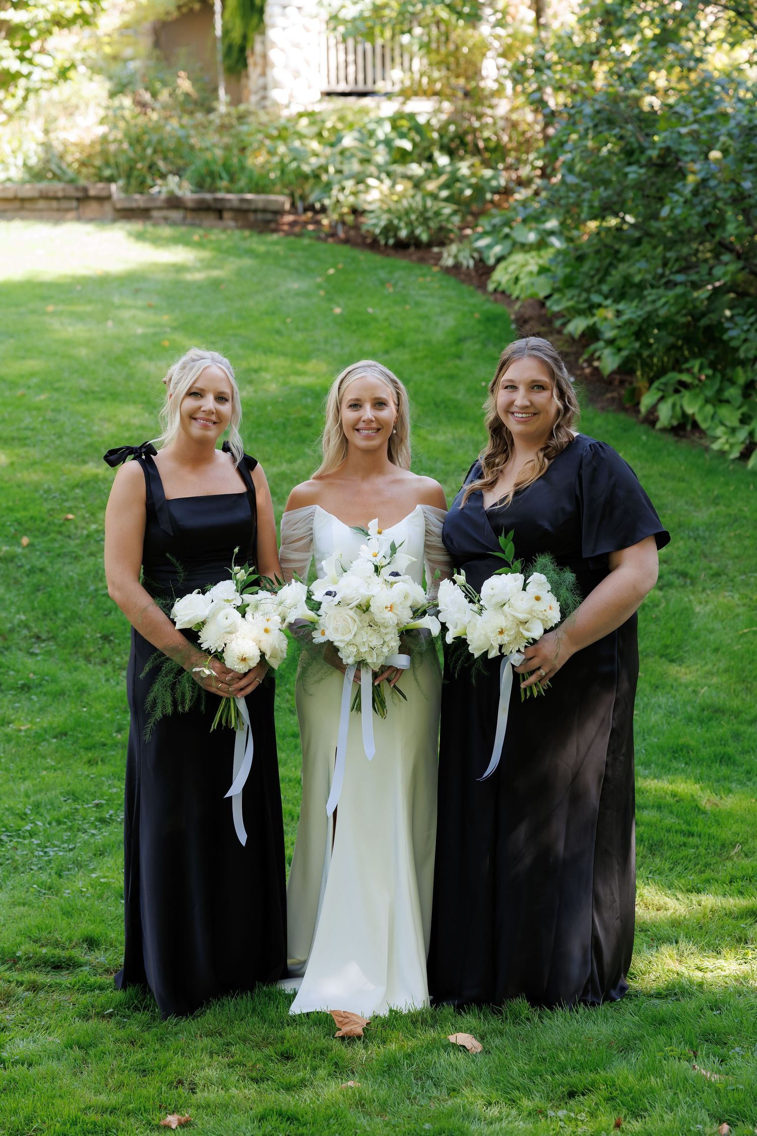 Bridesmaids in black dresses hold white bouquets while posing with bride in white.