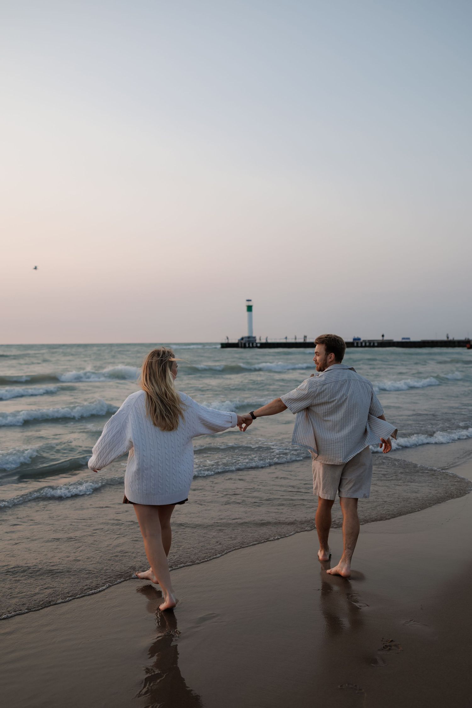Couple walking playfully along the beach at sunset near a lighthouse.
