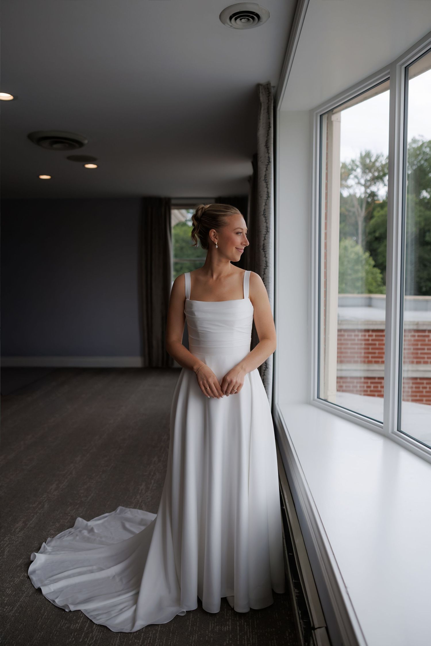 Bride in elegant white A-line wedding gown with train standing by large windows in soft natural light.