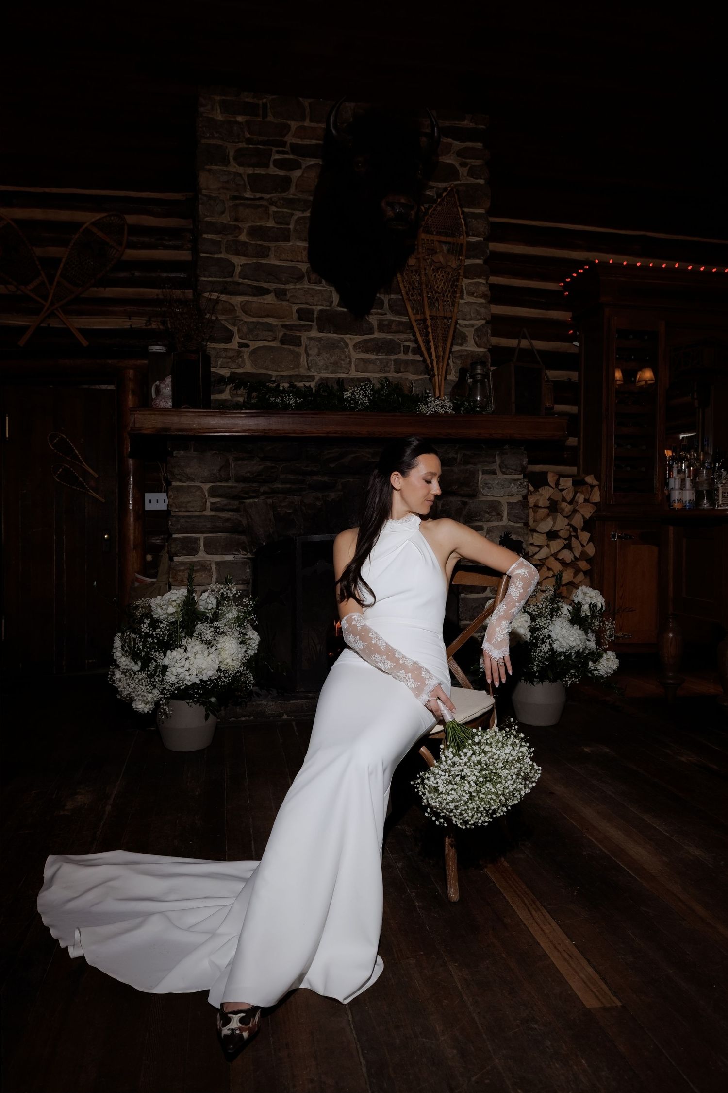Bride in white halter mermaid gown with lace gloves holds bouquet before rustic stone fireplace.