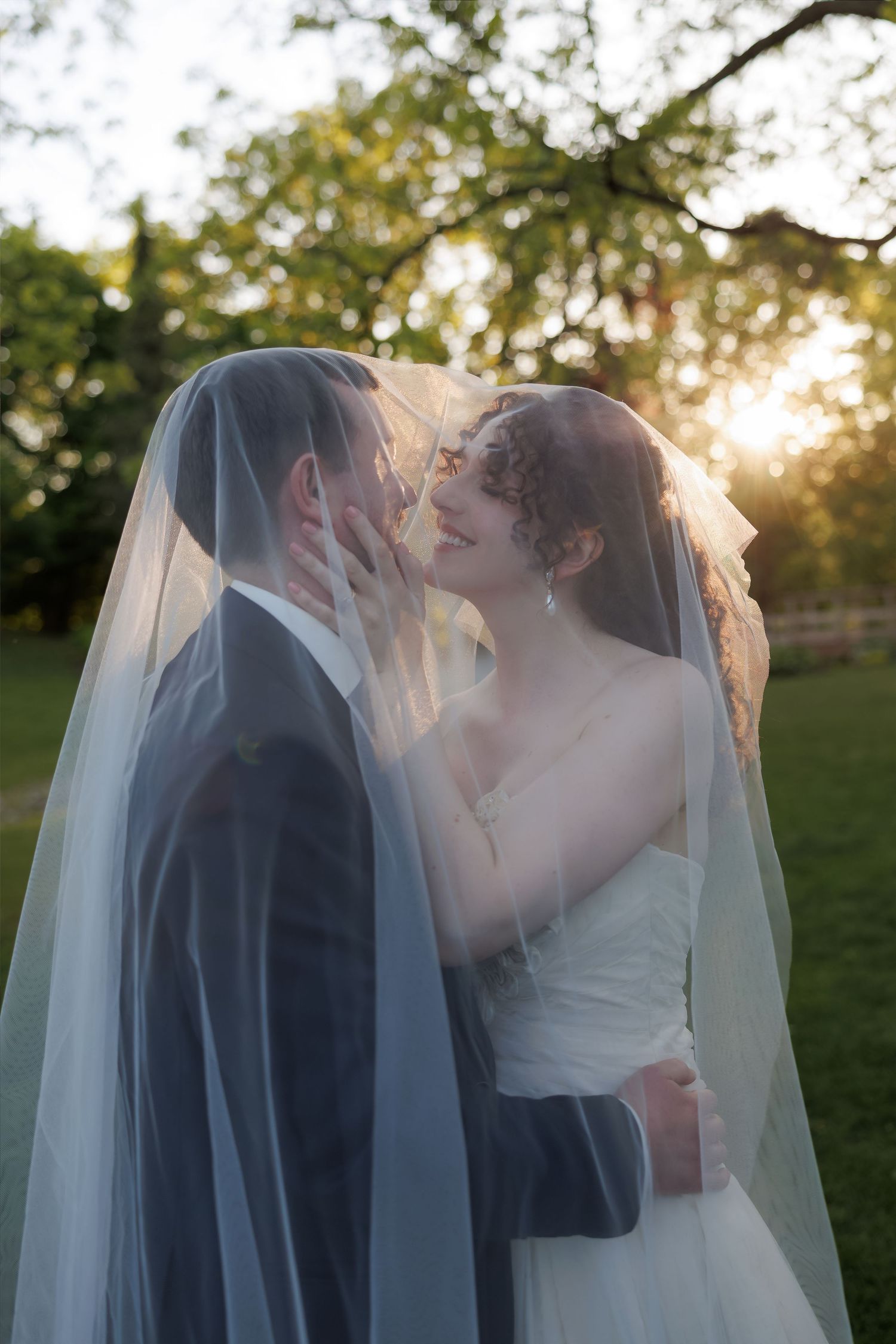 Bride and groom sharing a romantic kiss under a wedding veil at golden hour in a lush green outdoor setting.