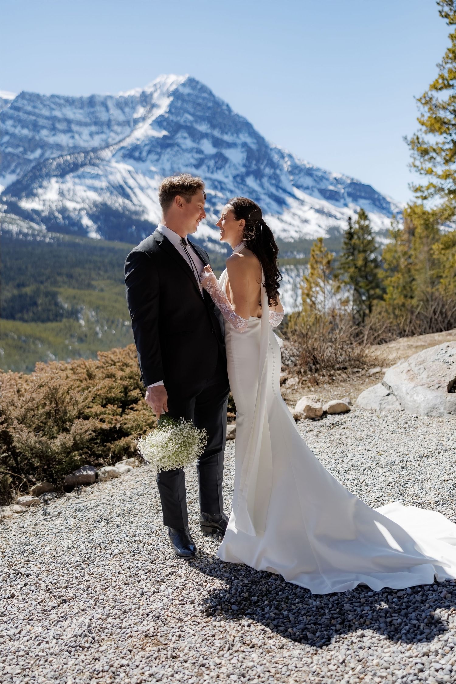 Bride and groom share romantic moment in white wedding attire with stunning snow-capped mountain scenery behind them.