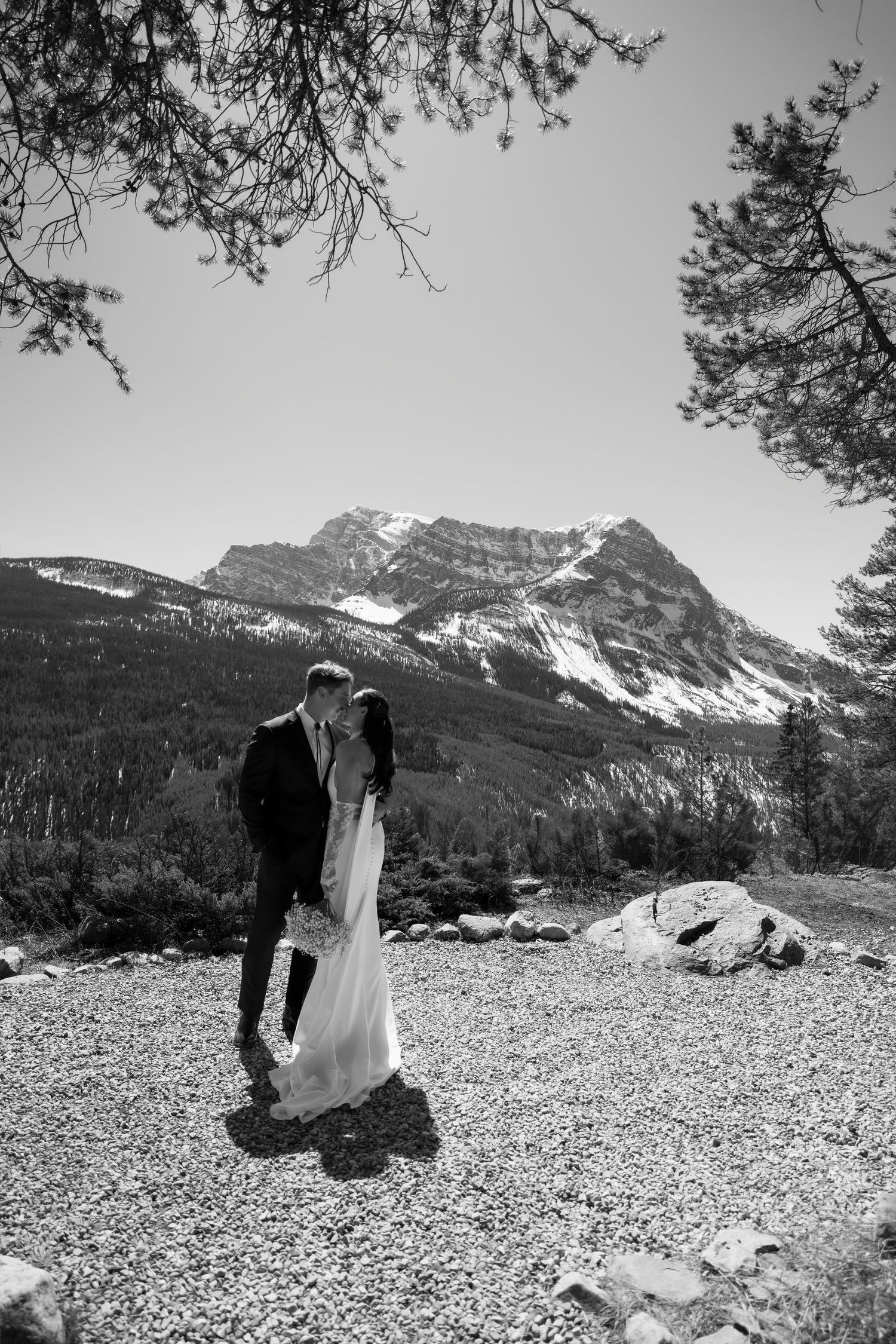Black and white photo of a couple kissing in front of a snow-capped mountain and pine trees on their wedding day.
