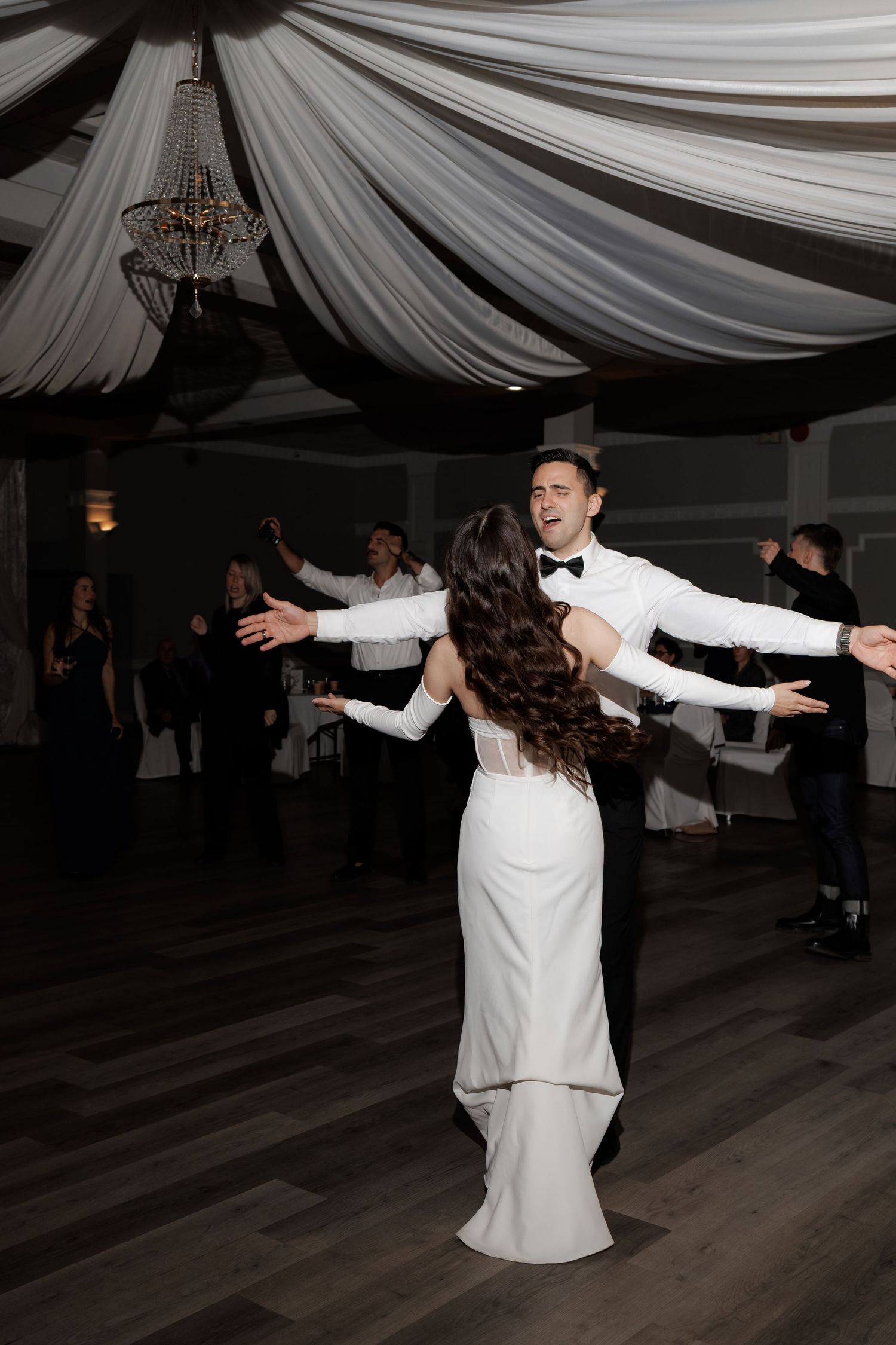 Wedding couple performs energetic dance moves during their reception under draped white fabric ceiling.