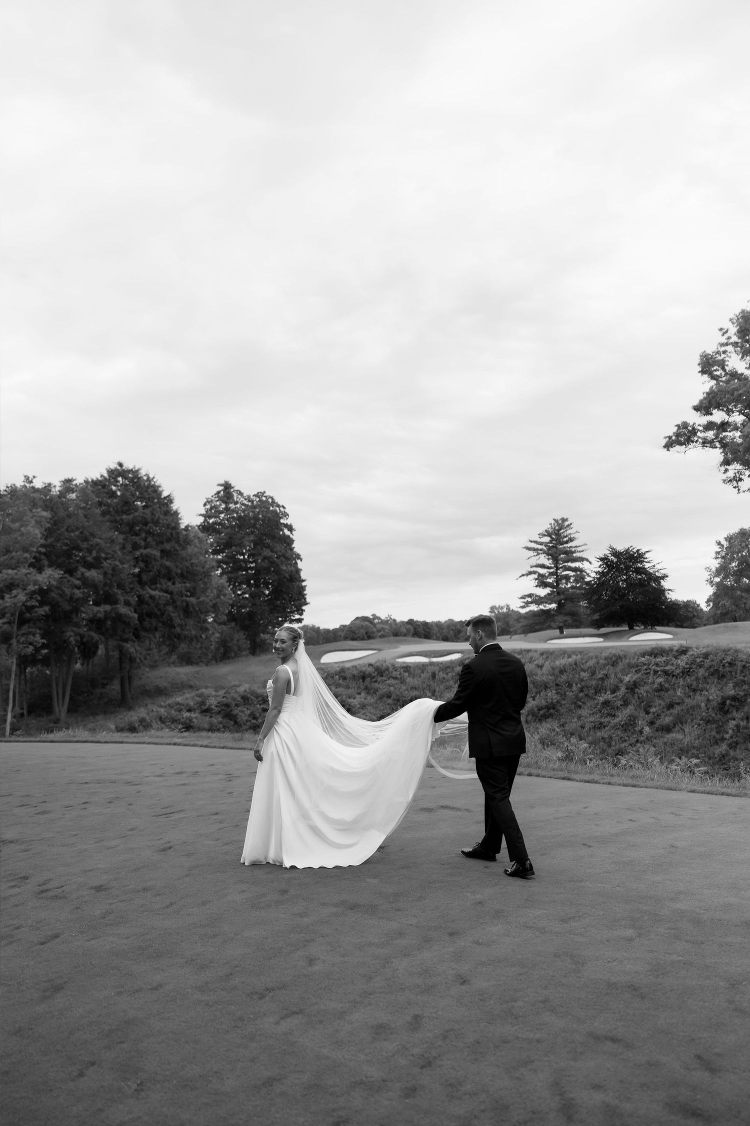 Black and white photo of bride and groom walking on golf course path, veil flowing in the wind.