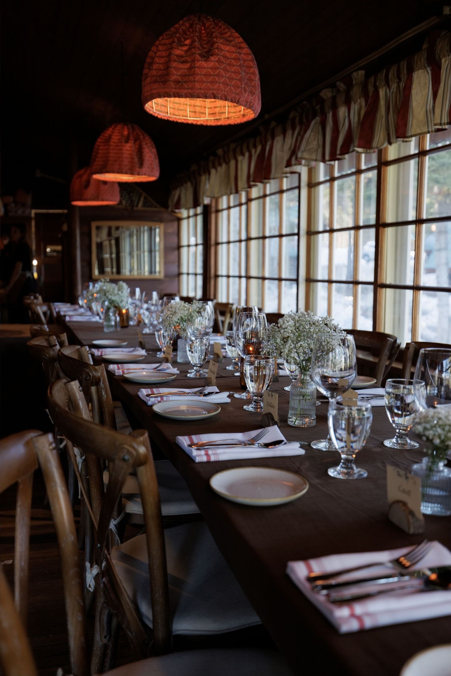 Elegant long dining table set with glassware, white floral centerpieces, and wicker pendant lights in a cozy restaurant.