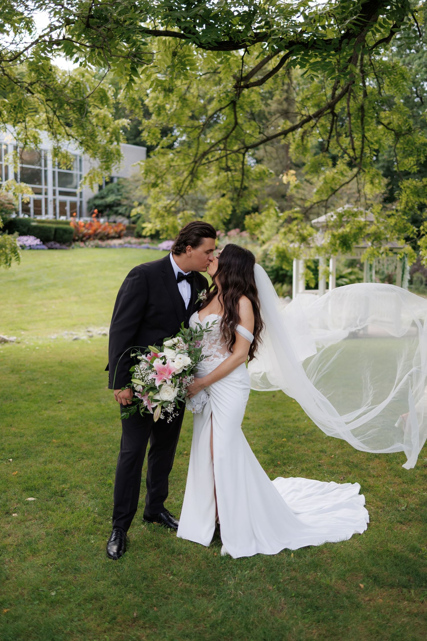 Bride and groom sharing a kiss outdoors, veil flowing in wind, white gown with train, bouquet of pink flowers, lush garden.