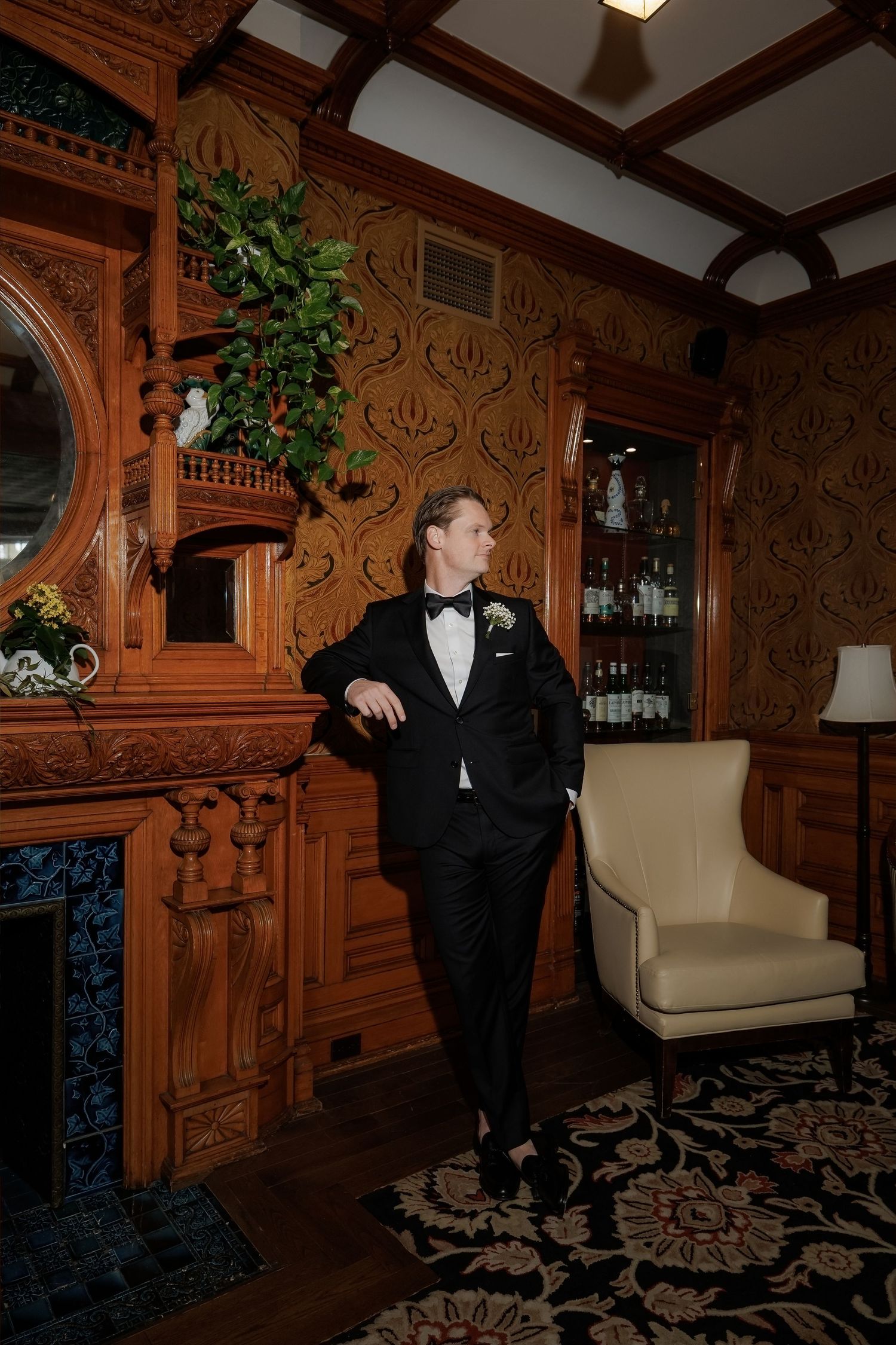 Dapper groom in black tuxedo with boutonniere leans against ornate wooden fireplace in vintage-style room.