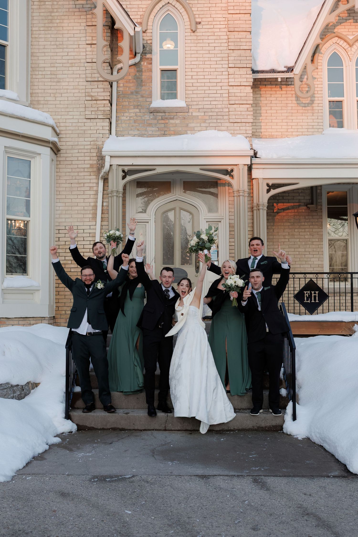 Wedding party celebrates on snowy steps of historic brick venue with bridesmaids in green dresses.