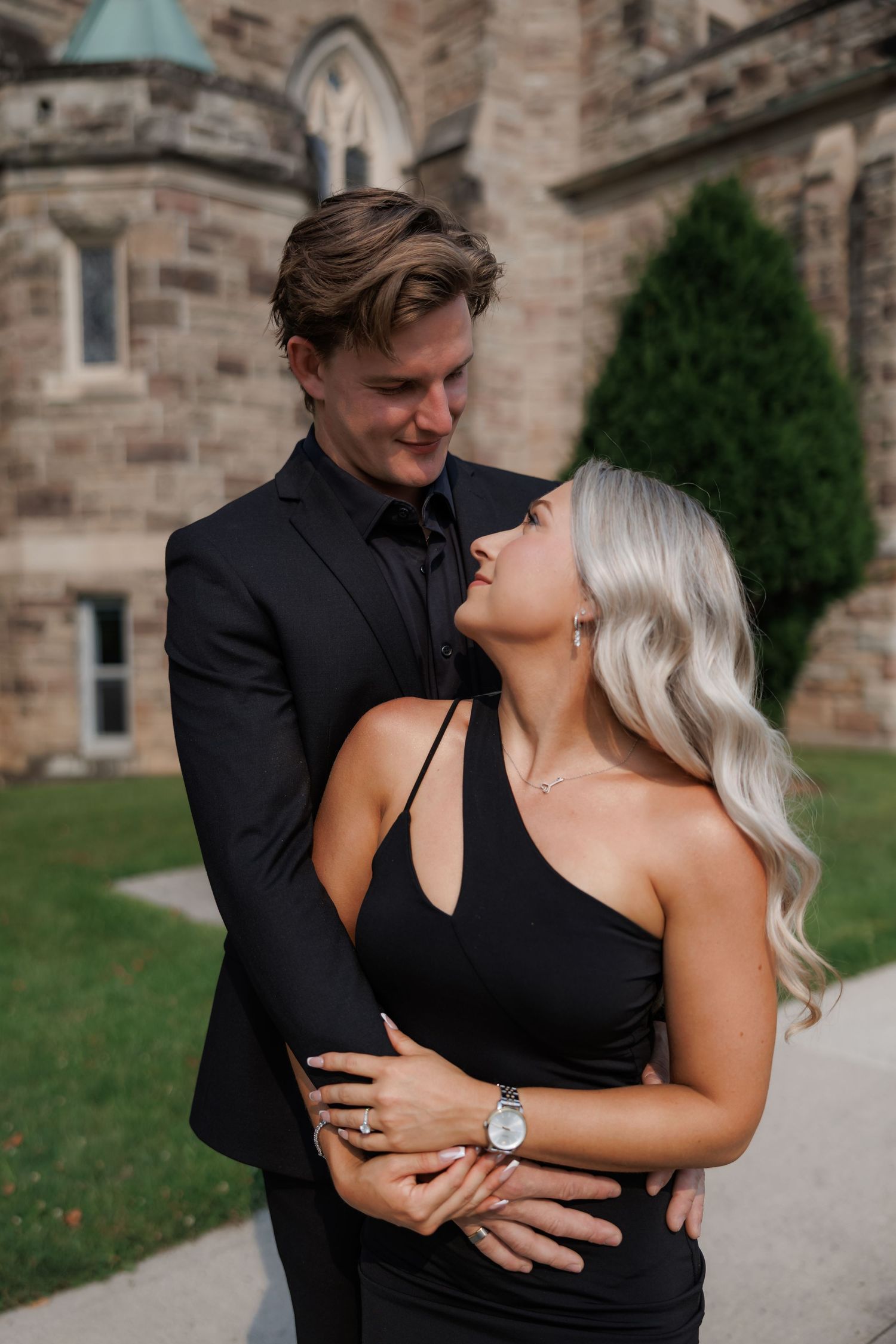 Couple in black formal wear embracing outside stone building.