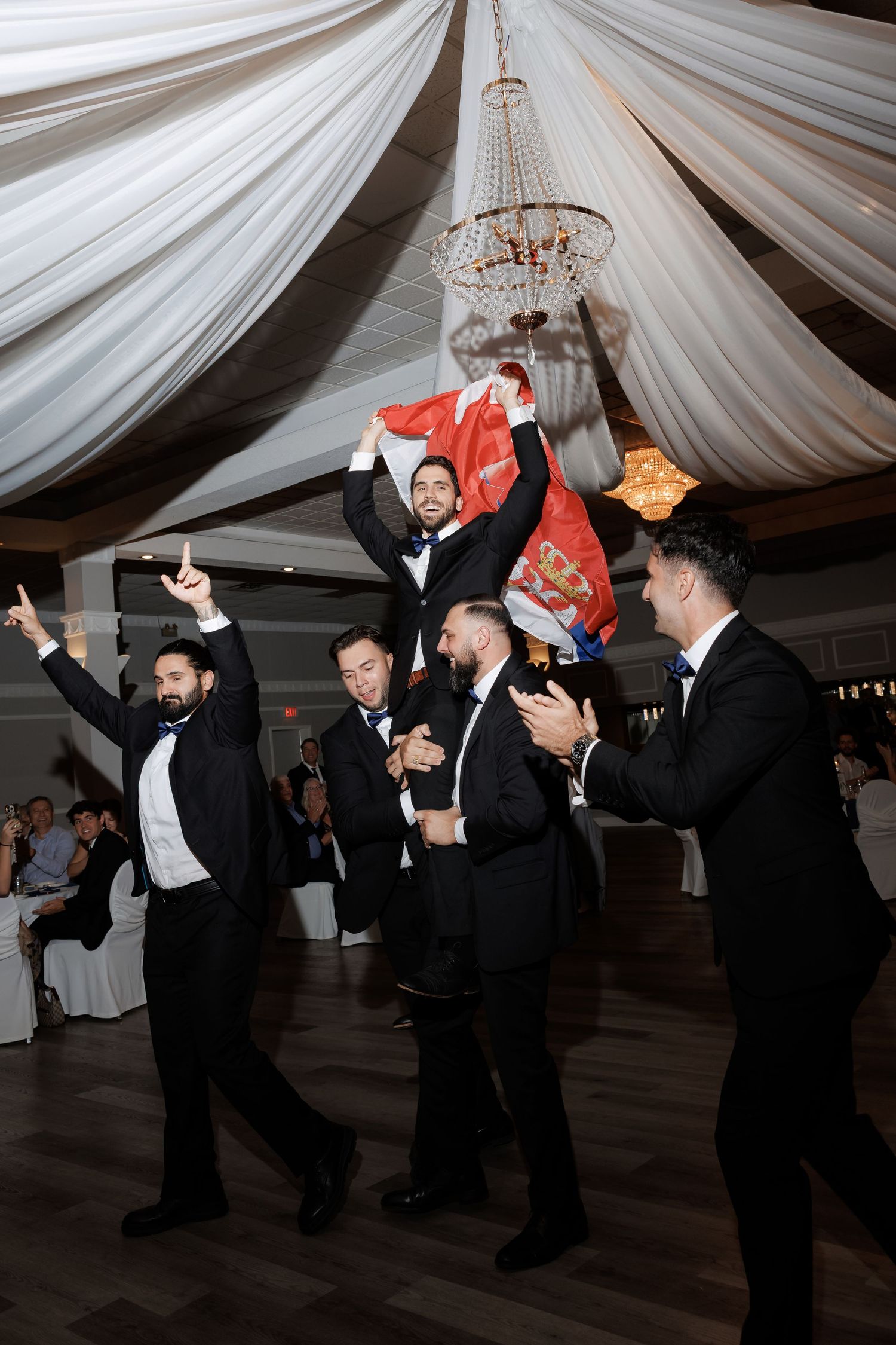 Wedding reception guests dancing energetically under white draped ceiling with chandeliers.