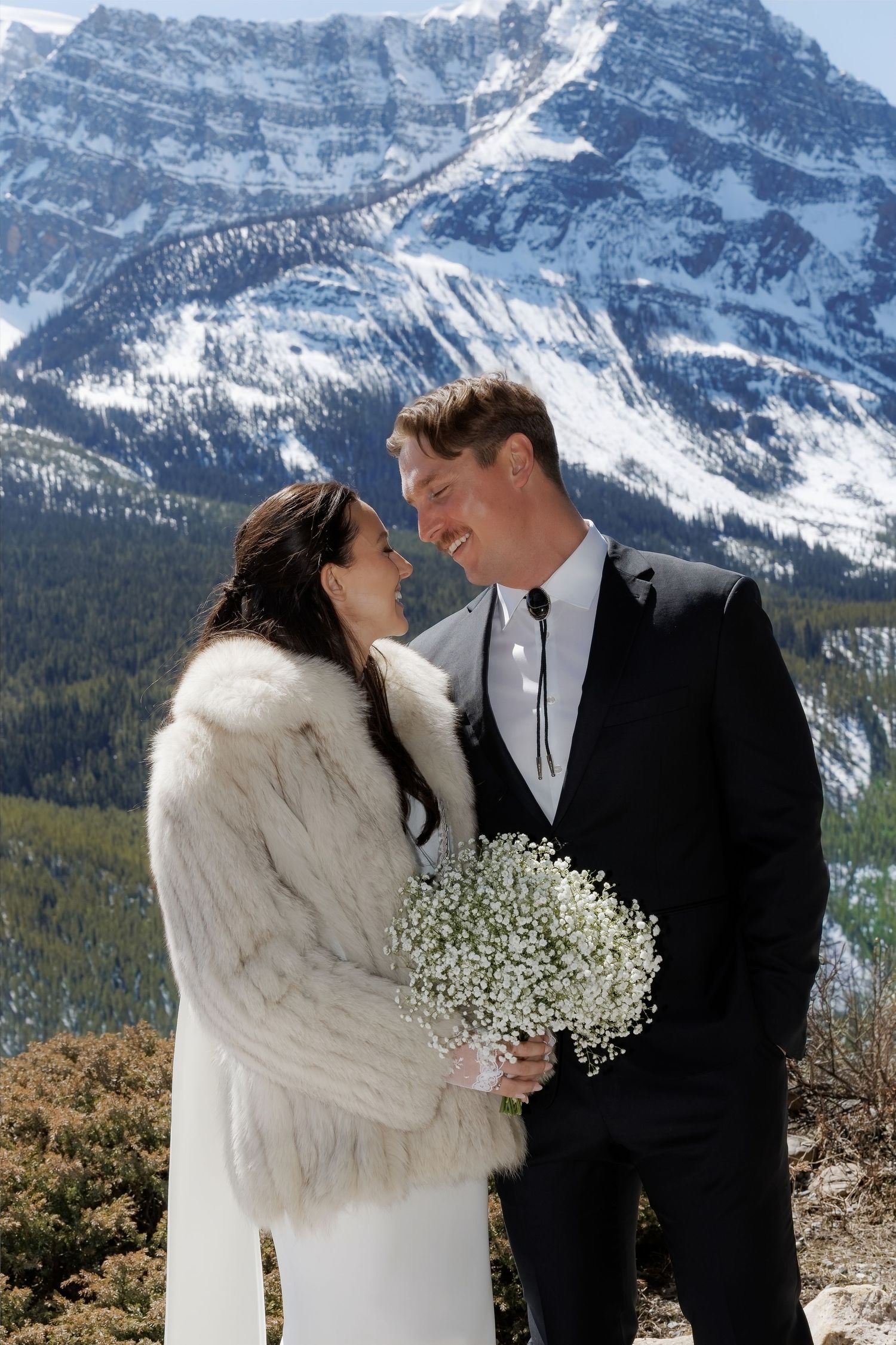 Bride and groom pose together with snowy mountain backdrop, she holds baby's breath bouquet wearing fur coat.