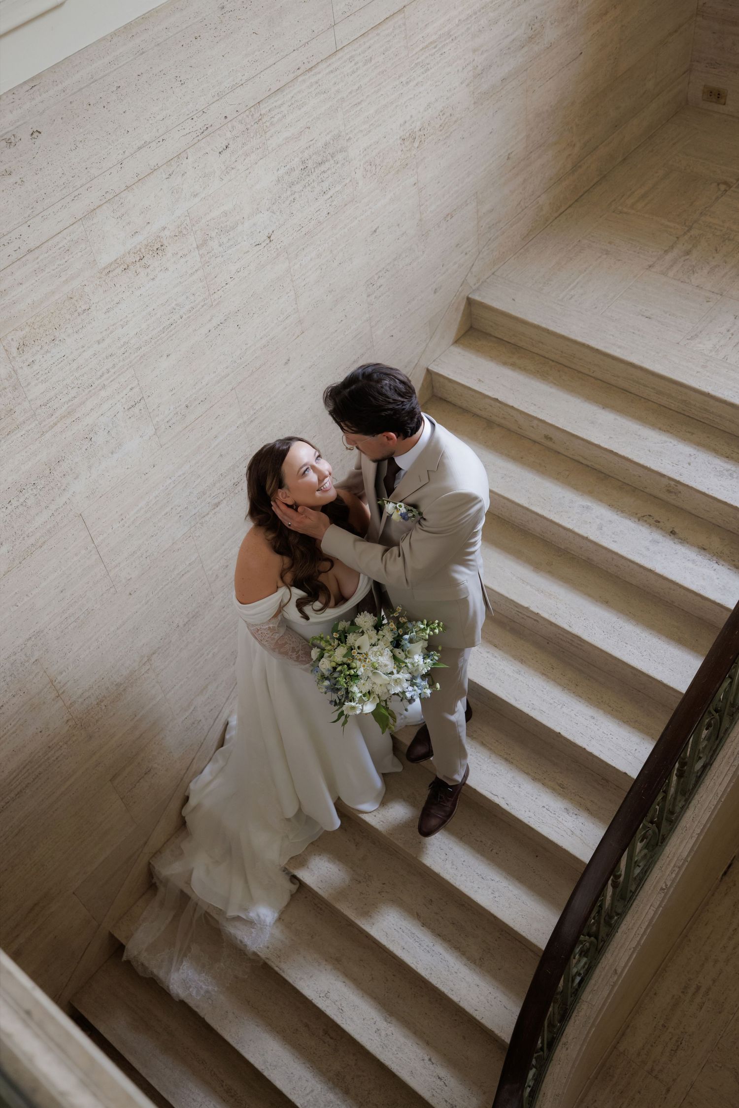 Aerial view of bride and groom embracing on elegant marble staircase, bride holding white floral bouquet.