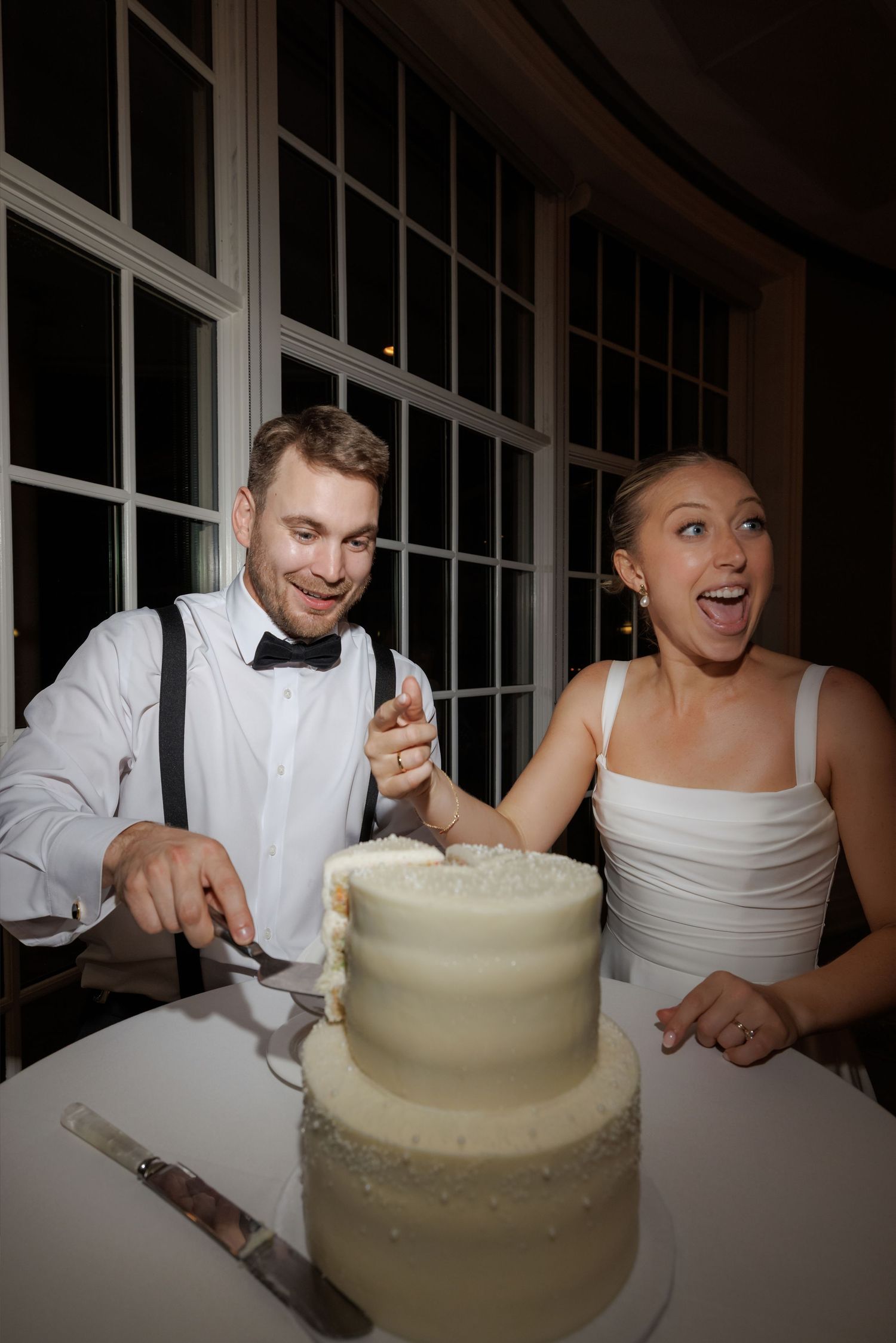 Excited bride and groom cutting white three-tier wedding cake at evening reception.