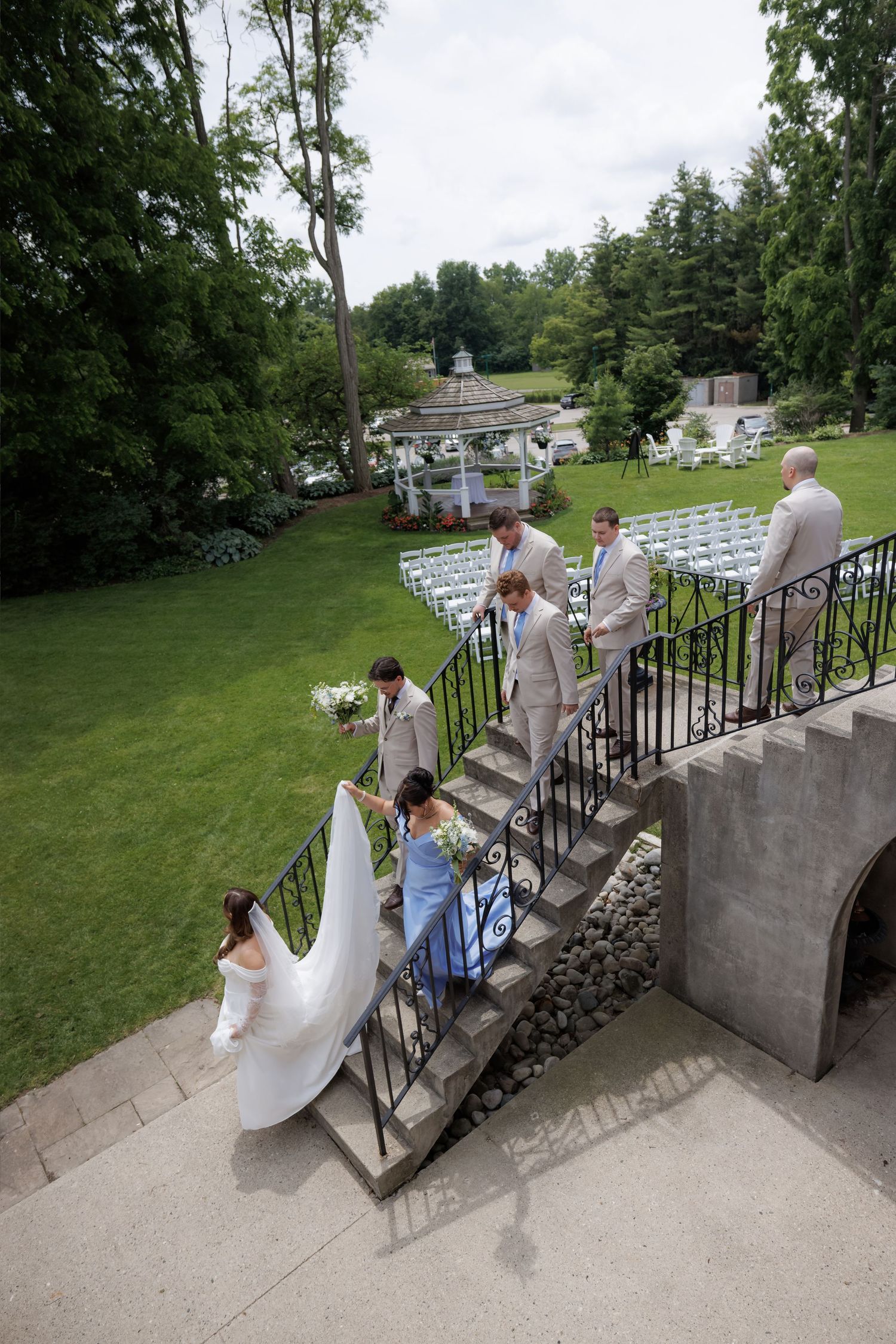 Bride in white gown with wedding party descending outdoor stone staircase to garden ceremony.