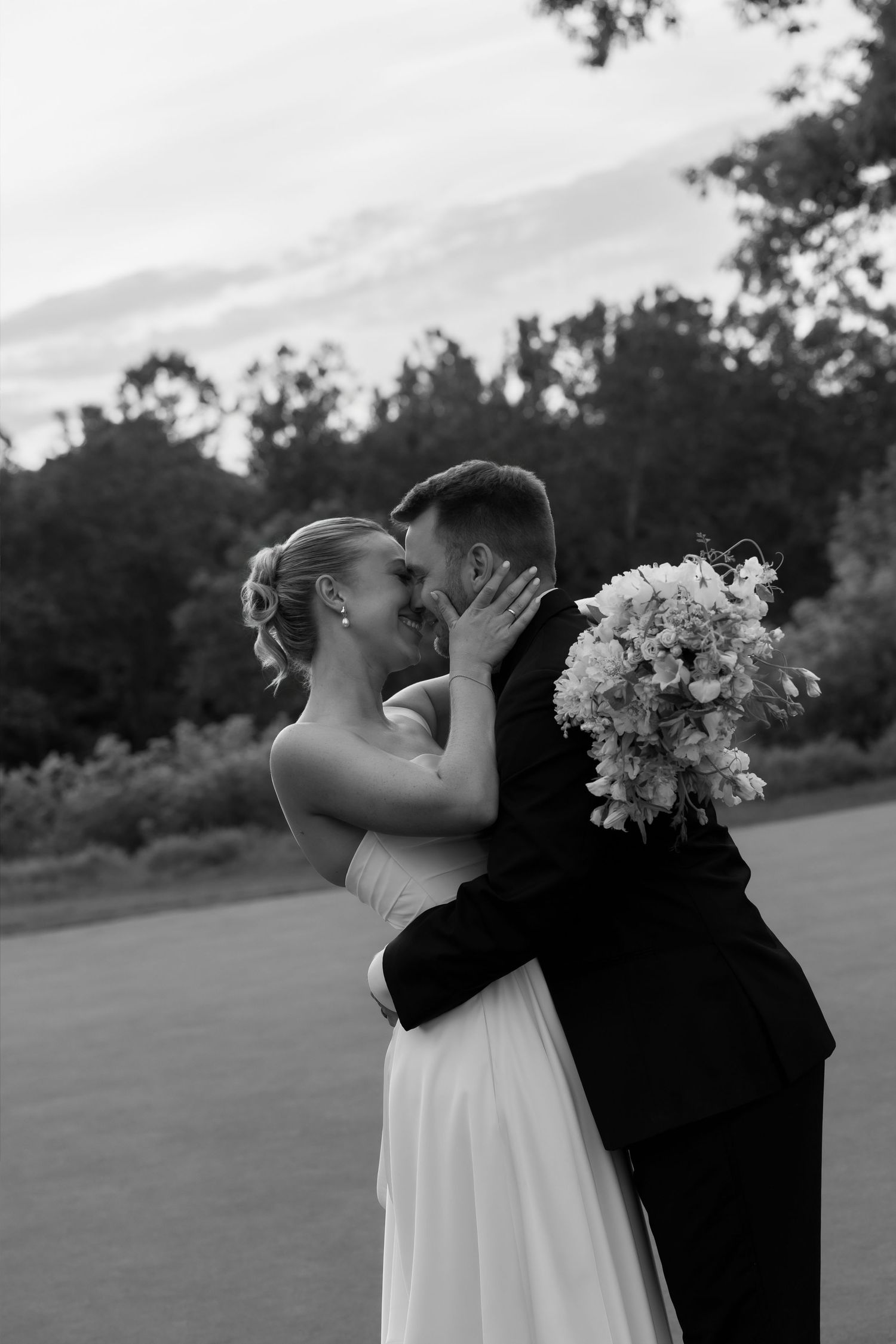 Black and white romantic photo of bride and groom embracing face to face holding wedding bouquet.