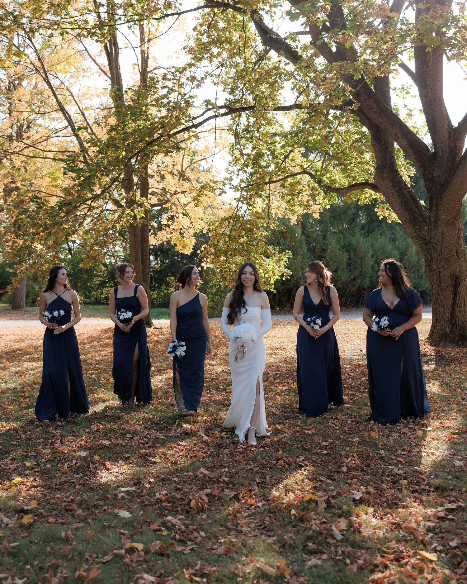 Wedding party poses under trees with golden autumn leaves on ground.