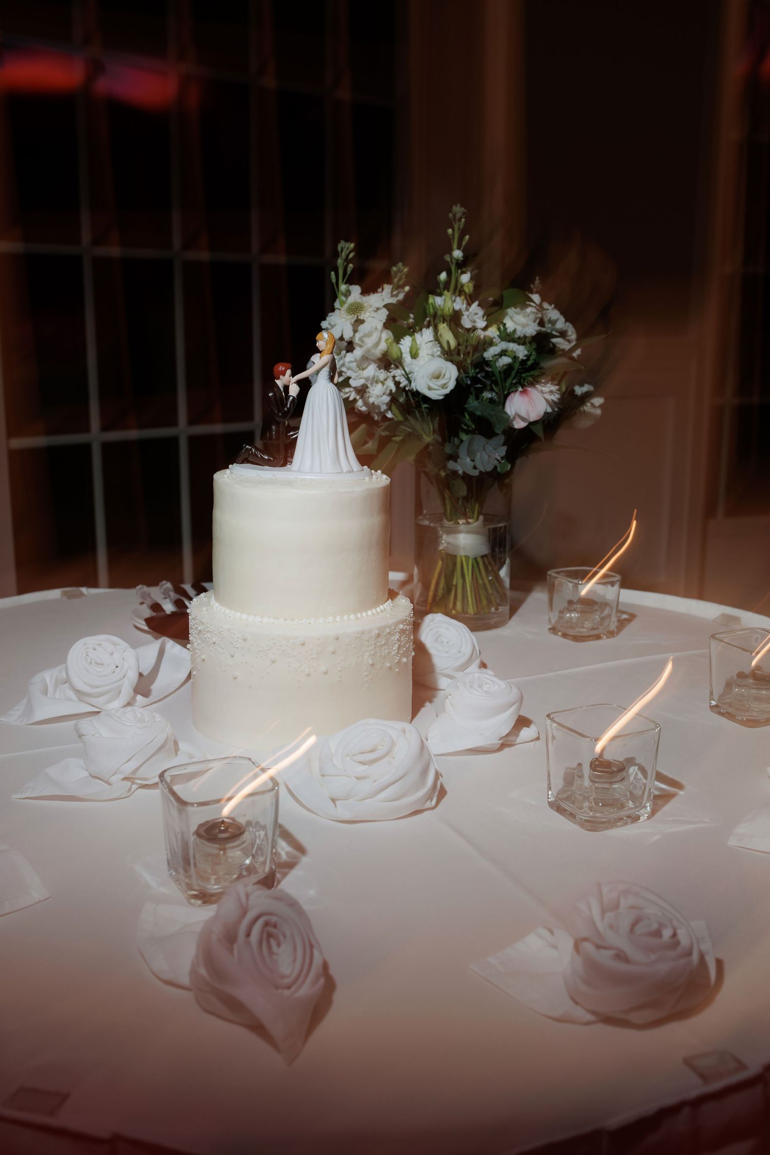 White wedding cake and floral centerpieces displayed on an illuminated table.