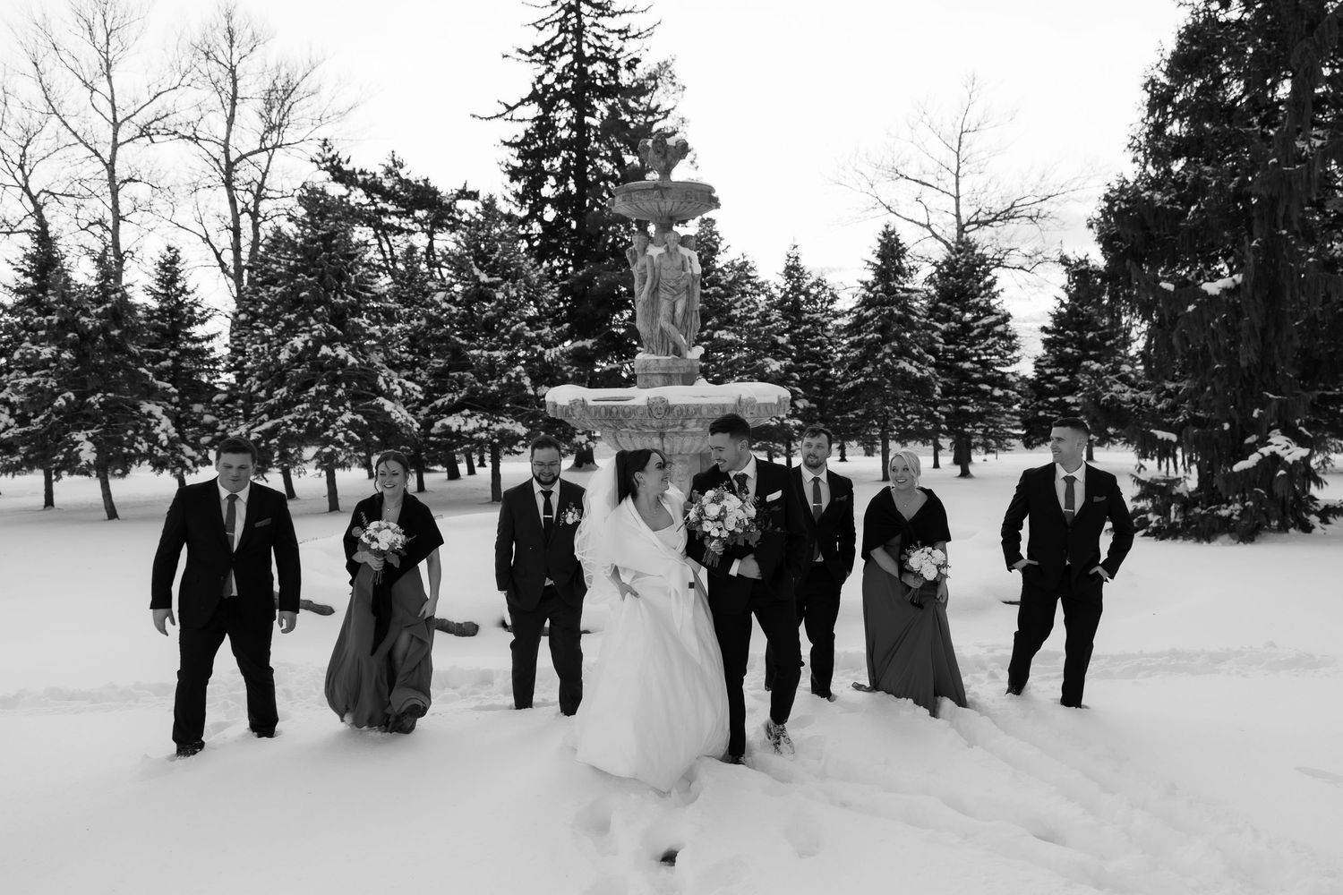 Wedding party posing in snowy winter landscape with evergreen trees and fountain in black and white photo.
