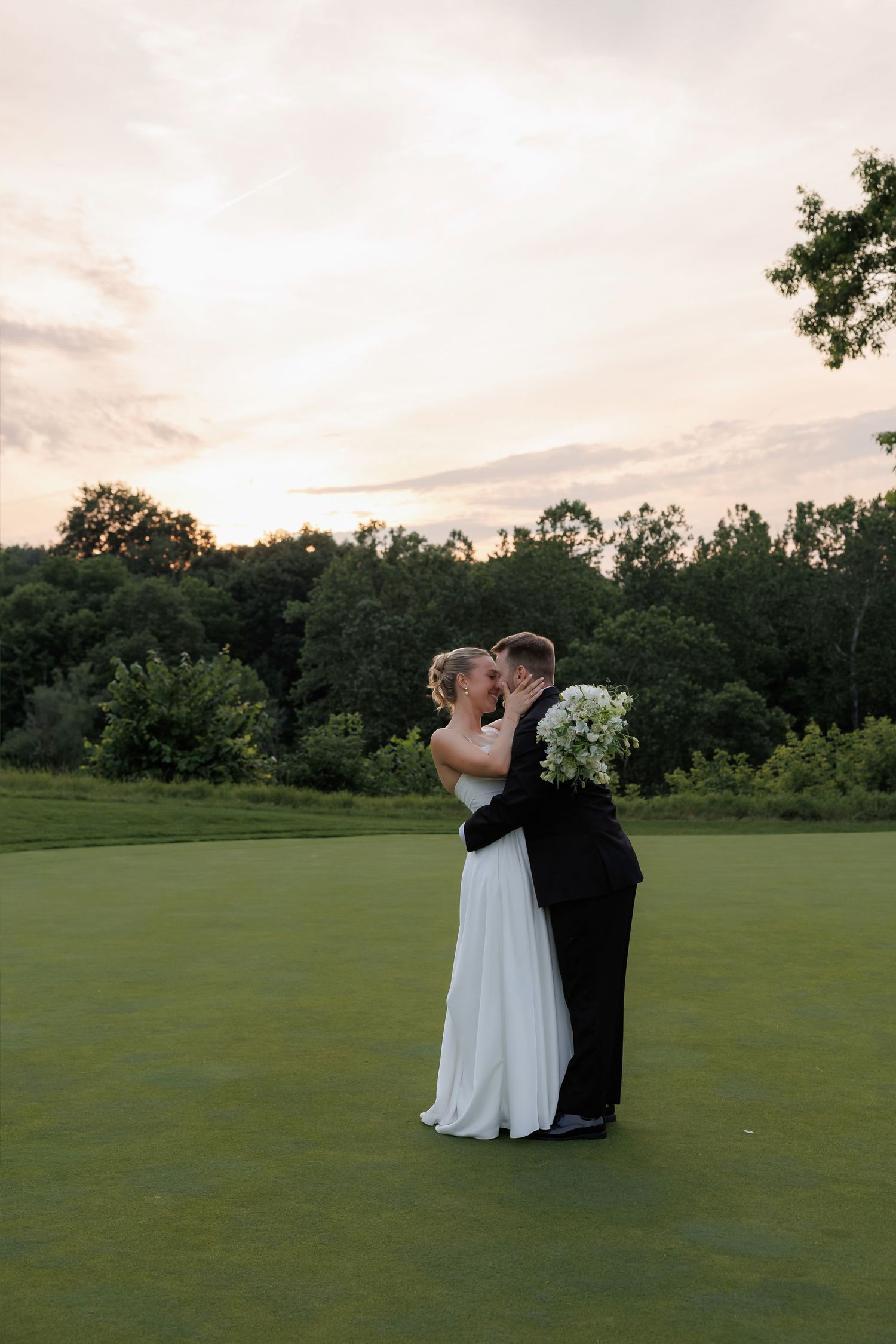 Bride and groom sharing romantic kiss on golf course green at sunset after wedding ceremony.