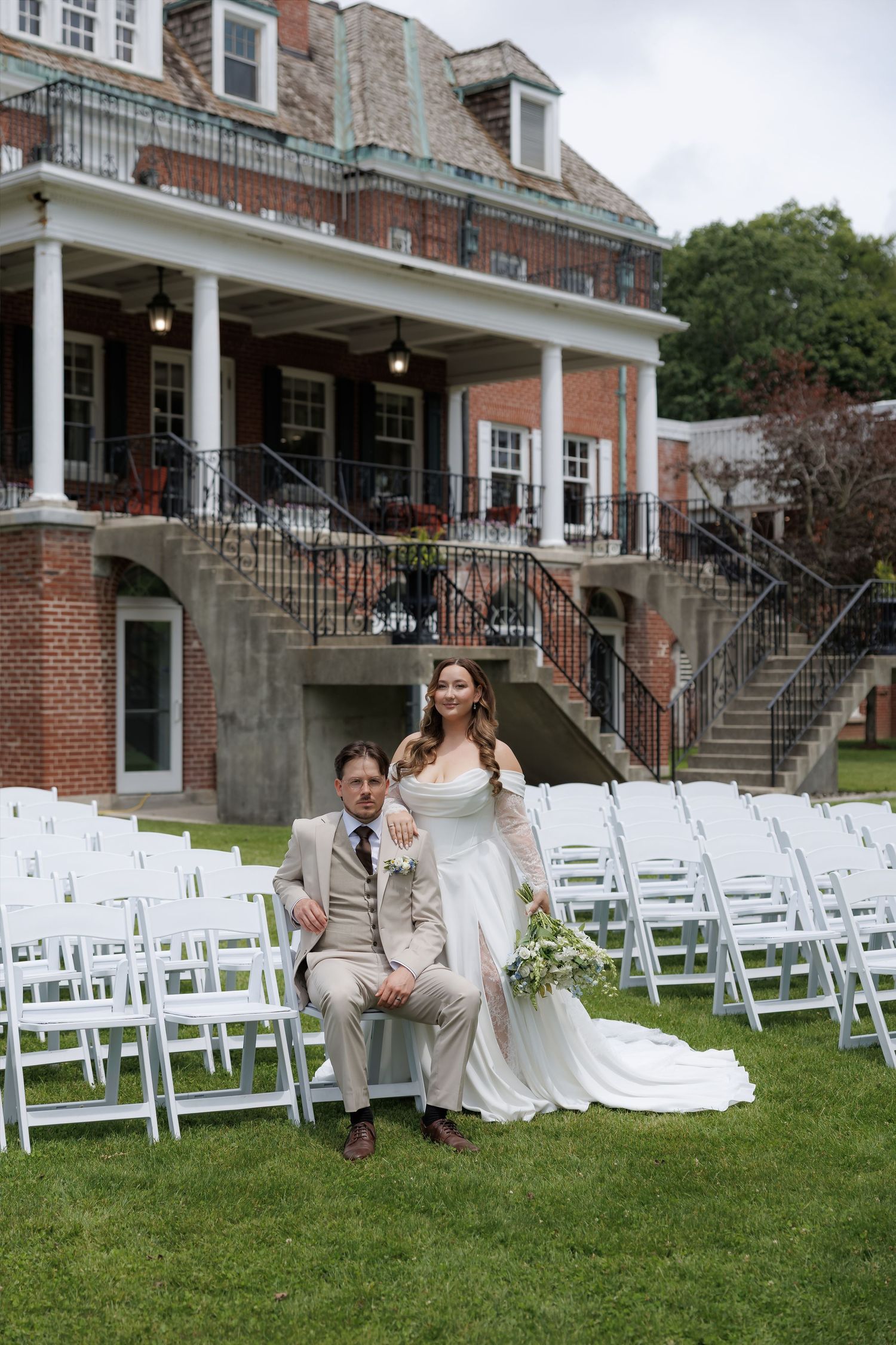 Bride and groom pose together on lawn chairs before a brick manor house with white ceremony chairs arranged behind them.