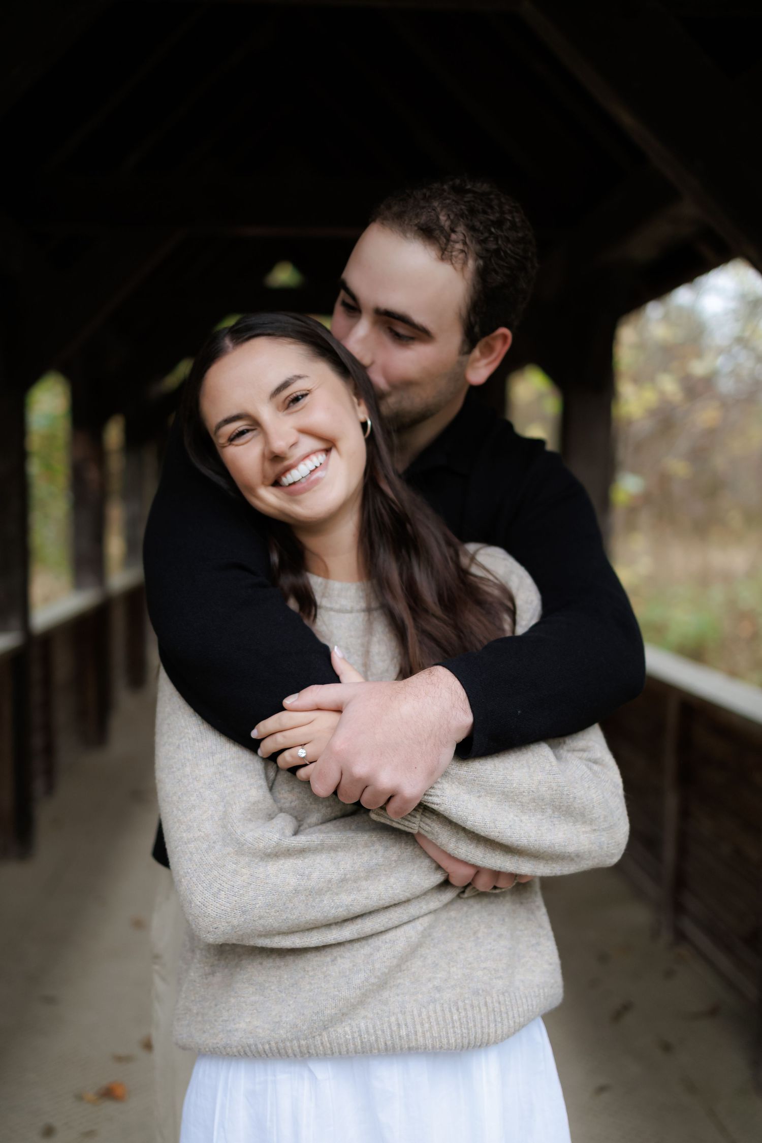 A couple shares a tender embrace in a rustic covered walkway, with the person in black hugging from behind.