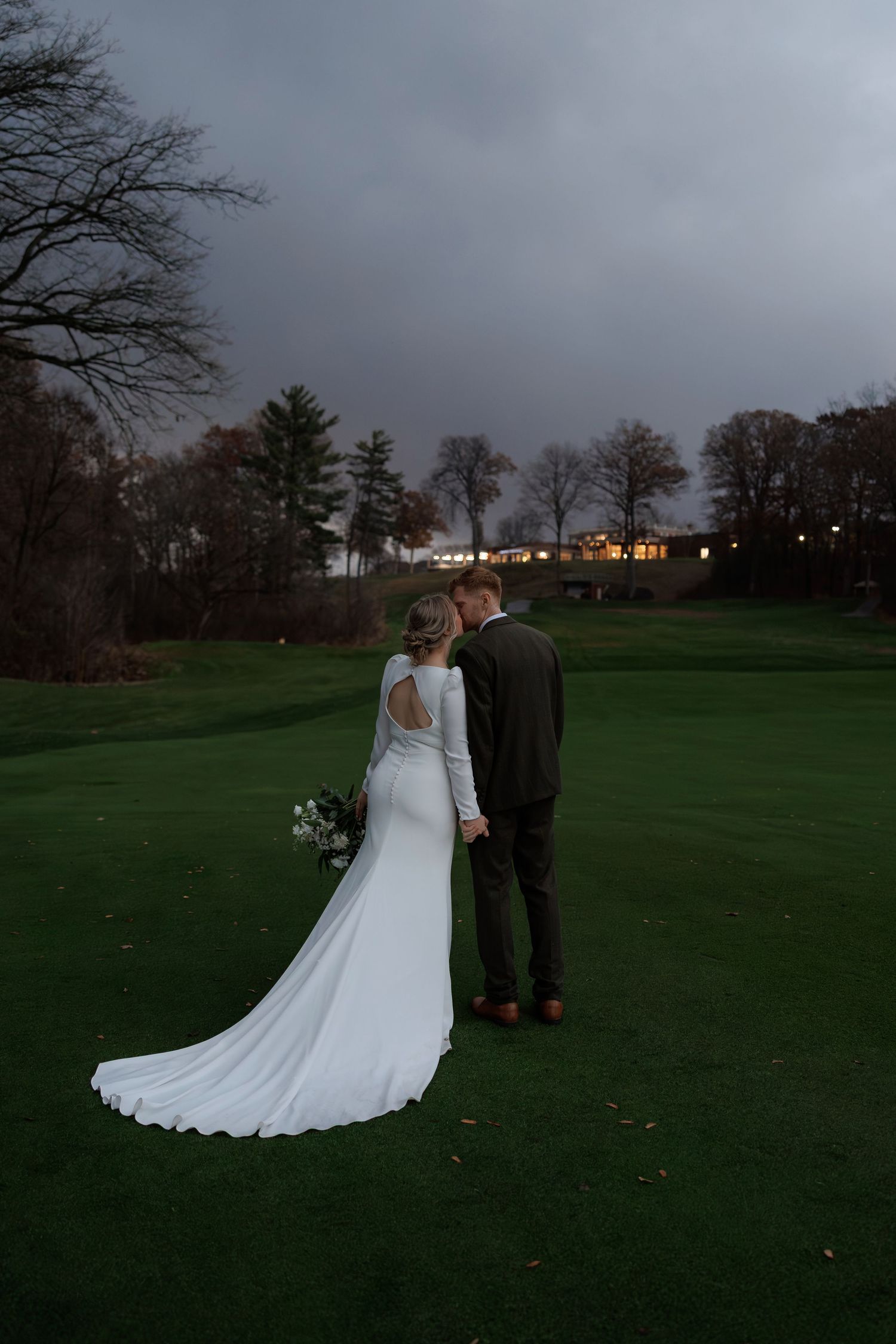 A couple in wedding attire stands on a green lawn at dusk with bare trees and buildings visible in the background.