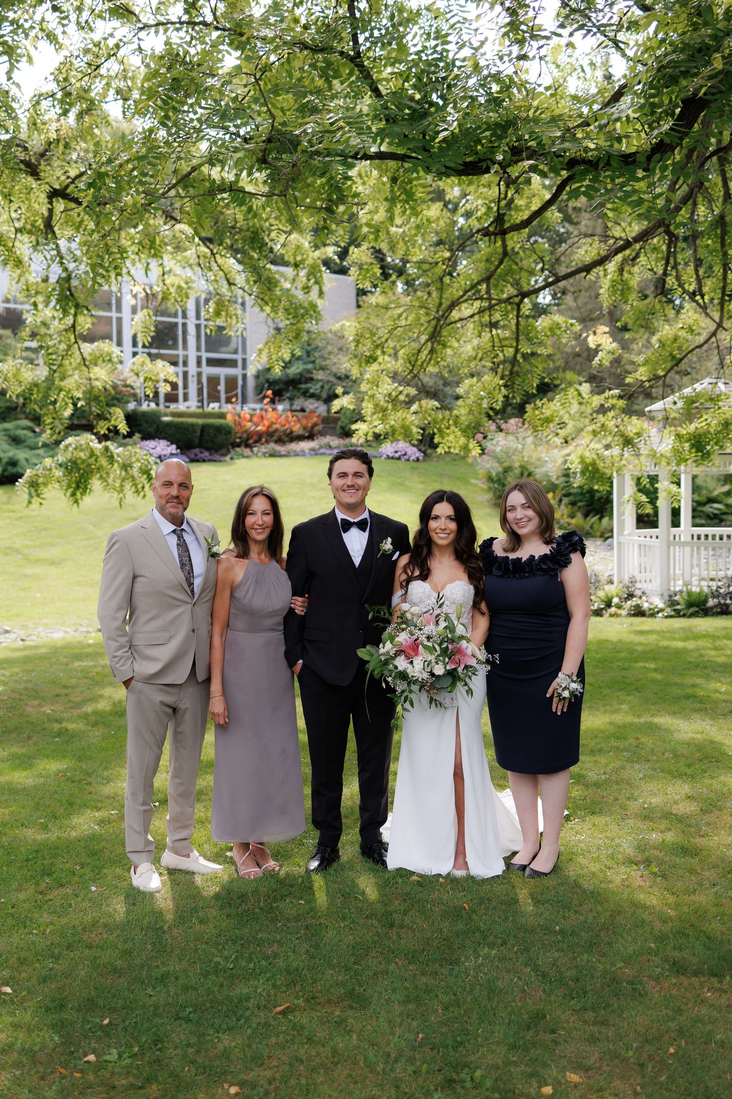 Wedding party poses for formal group photos on a green lawn with trees in the background.