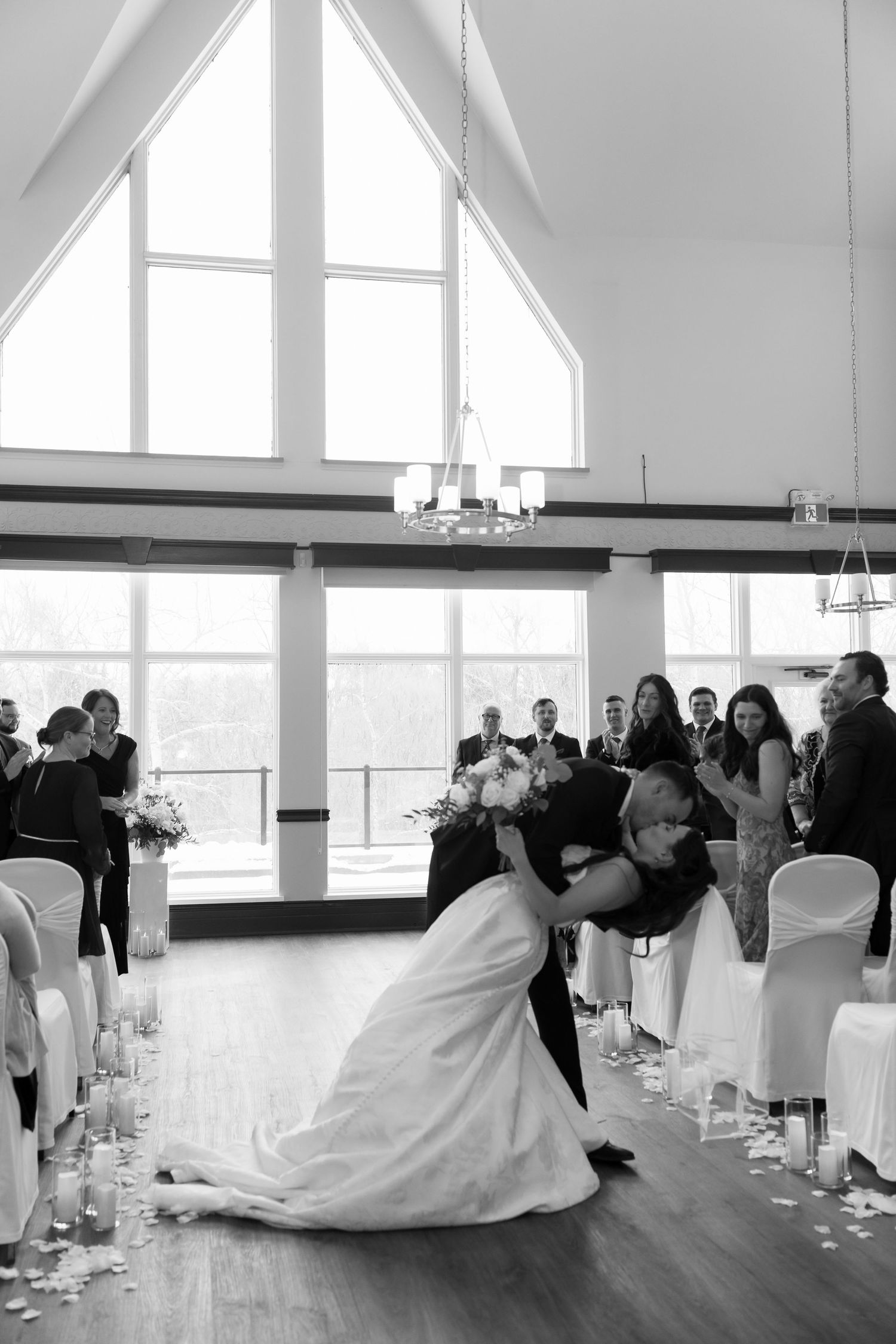 Groom dips bride for dramatic kiss in chapel aisle surrounded by cheering wedding guests.