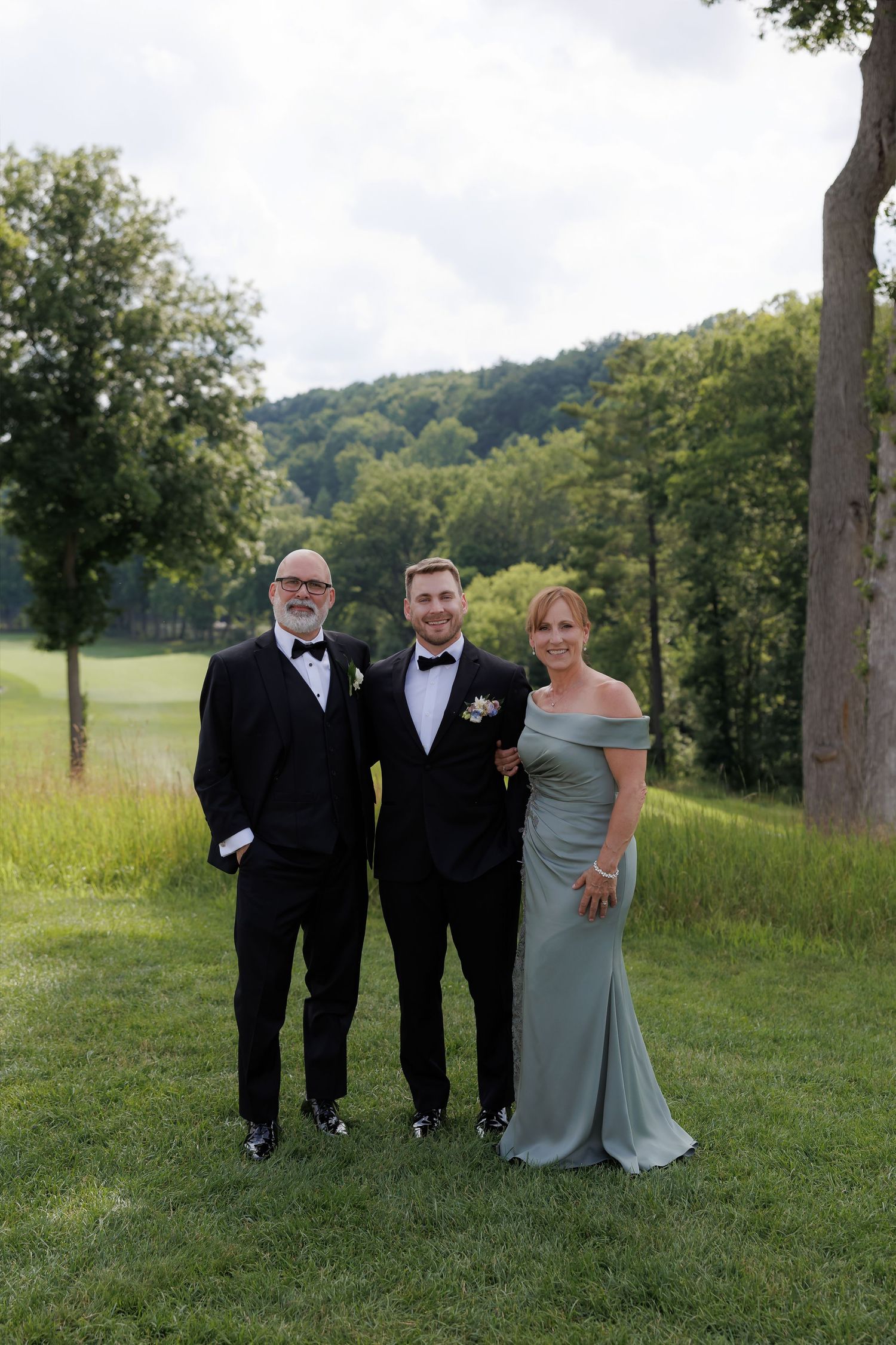 Groom poses with his parents on a lush green golf course, mother wearing a sage off-shoulder gown.