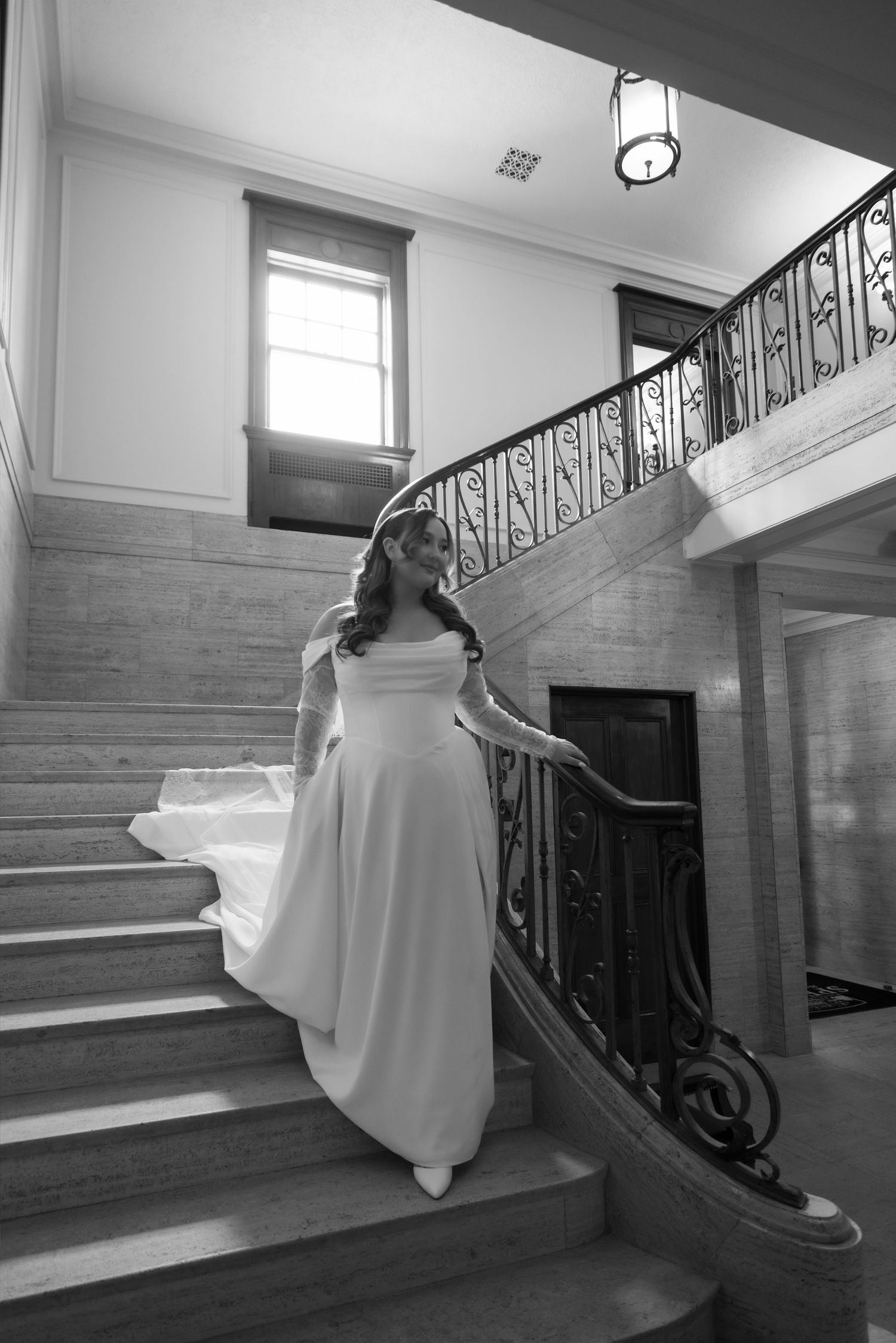 Elegant bride in white gown descending ornate curved staircase in black and white wedding photo.