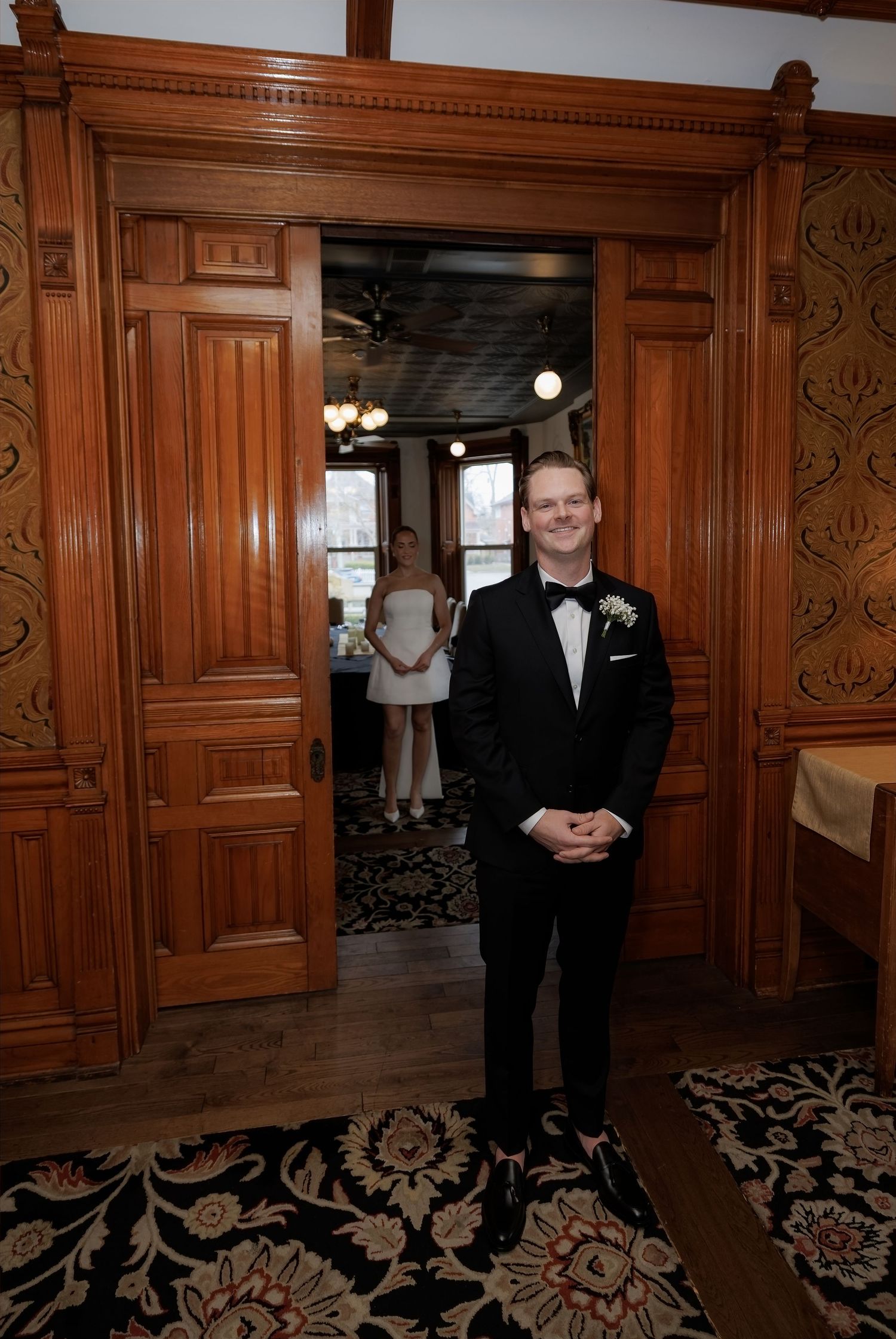 Groom in black tuxedo stands at ornate wooden doorway during first look moment with bride in background.