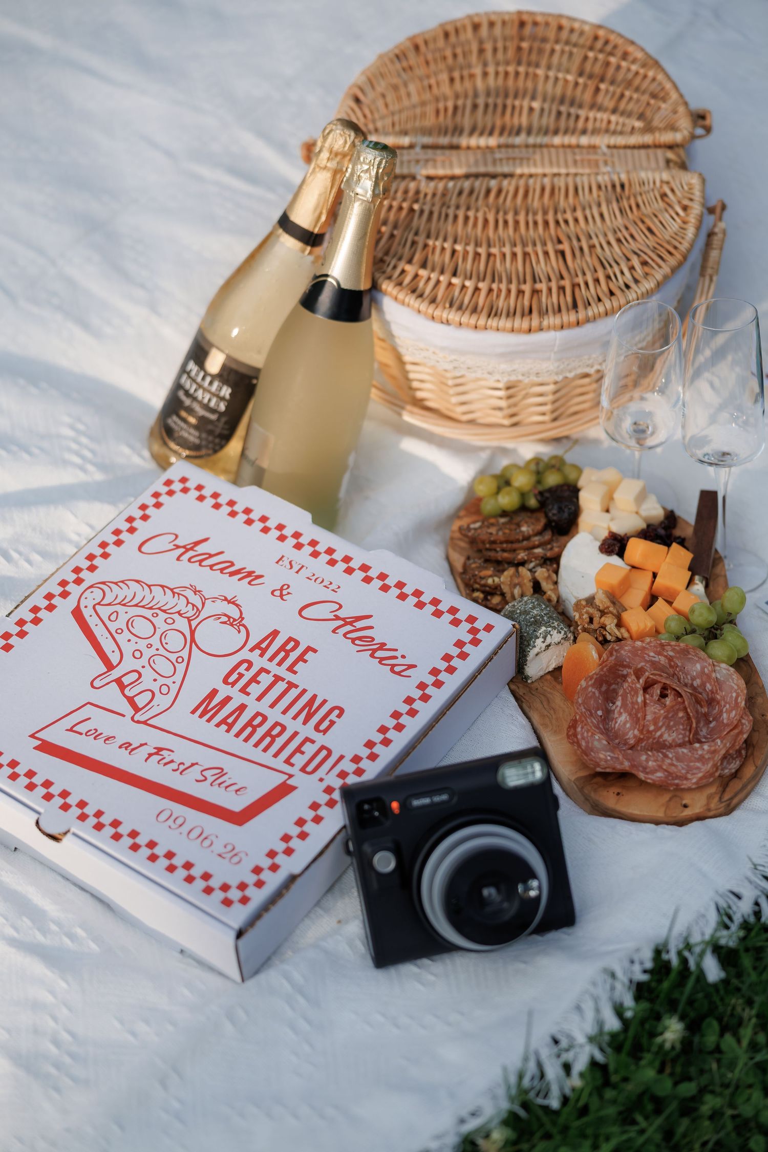 Picnic setup with wicker basket, food, and red checkered takeout boxes on a blanket.